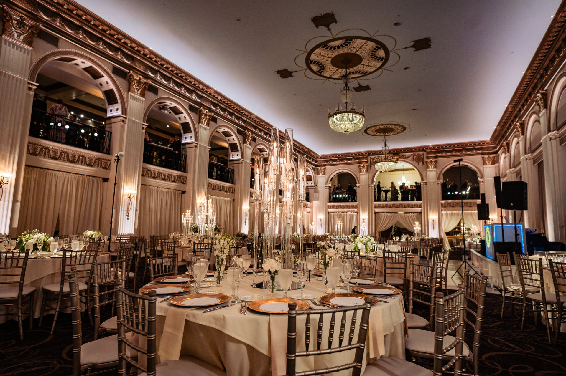 Elegant ballroom wedding reception with round tables set with gold-rimmed plates, white floral centerpieces, tall glass candle displays, and gold chiavari chairs beneath ornate arches and crystal chandeliers at The Ben.