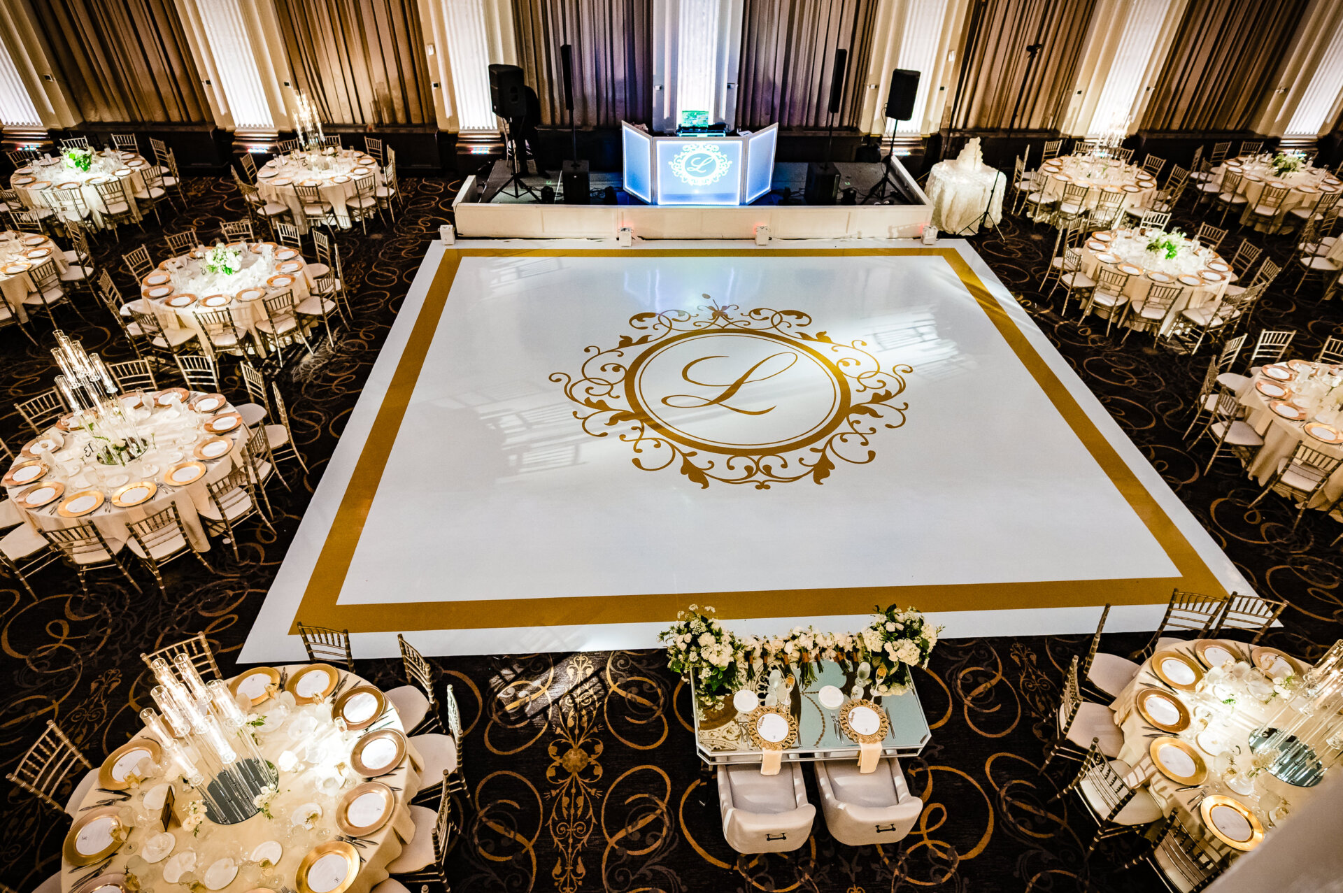 Overhead view of an elegant ballroom wedding reception featuring a custom white dance floor with gold monogram, surrounded by round tables with gold-rimmed place settings, white floral centerpieces, and gold chiavari chairs at The Ben.