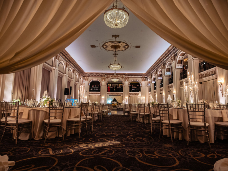 Elegant ballroom reception setup featuring round tables with white floral centerpieces, gold chiavari chairs, tall glass candles, and crystal chandeliers, framed by soft draped curtains at The Ben wedding venue.