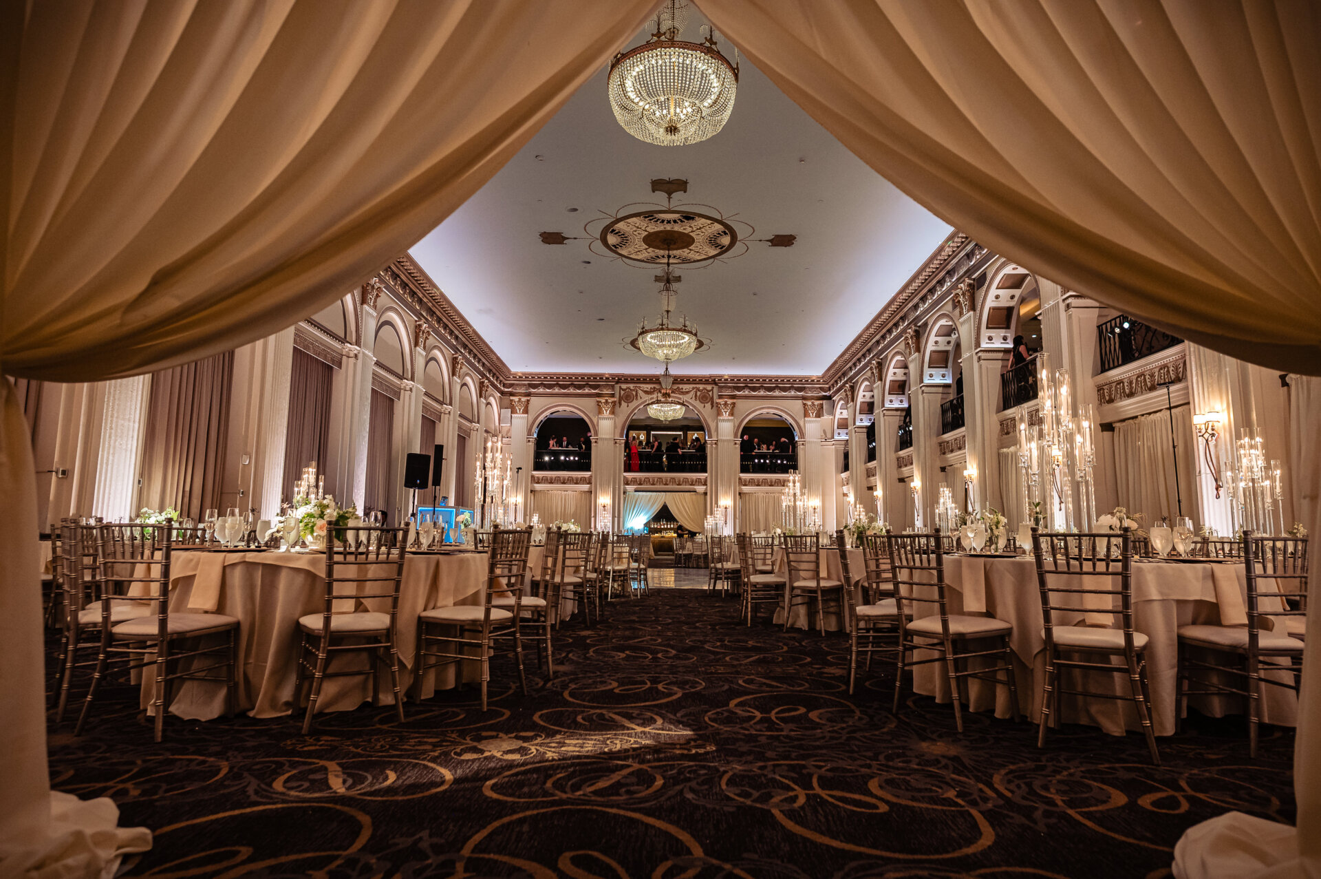 Elegant ballroom reception setup featuring round tables with white floral centerpieces, gold chiavari chairs, tall glass candles, and crystal chandeliers, framed by soft draped curtains at The Ben wedding venue.