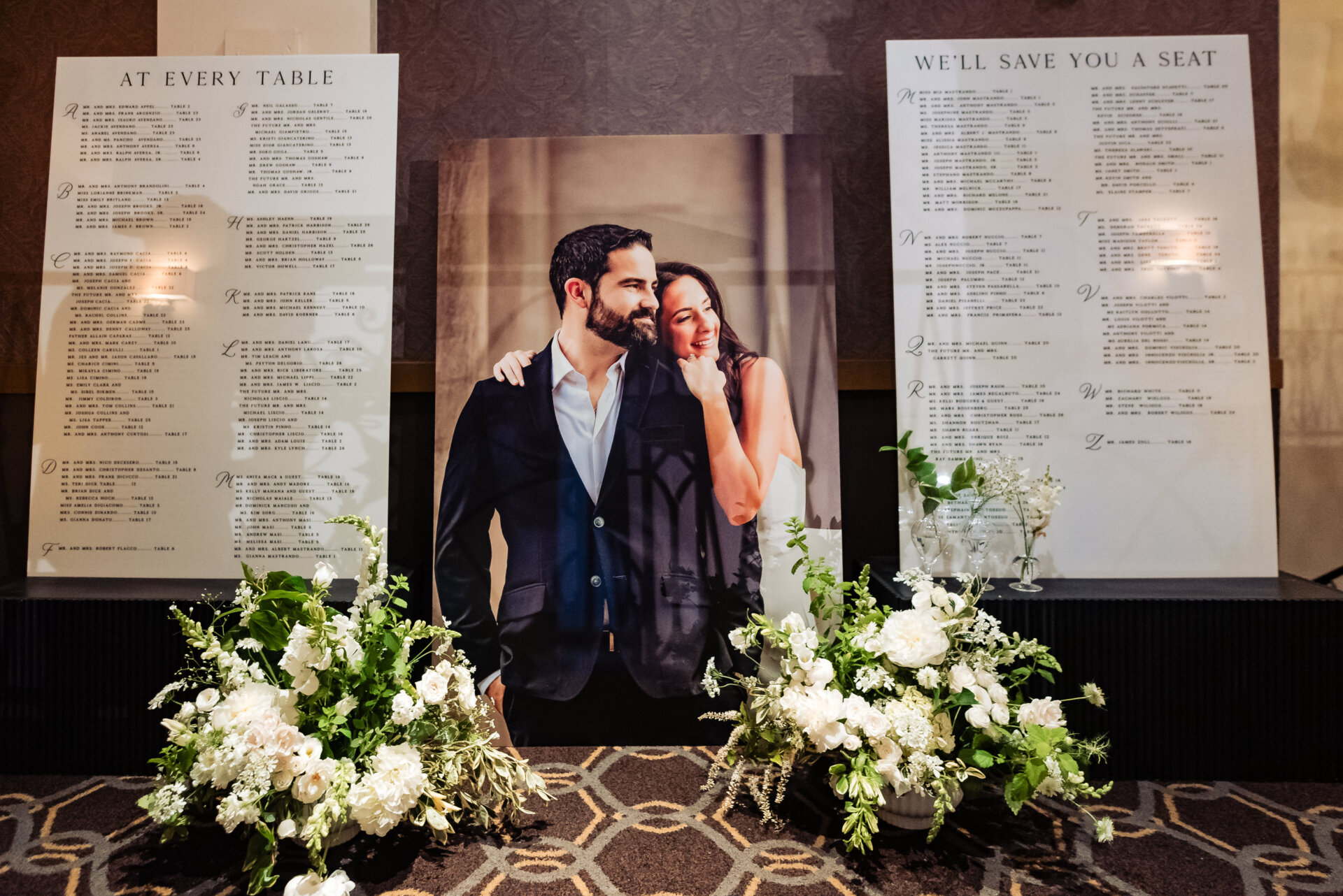 Wedding seating chart display featuring a large portrait of the couple, flanked by guest lists and white floral arrangements.