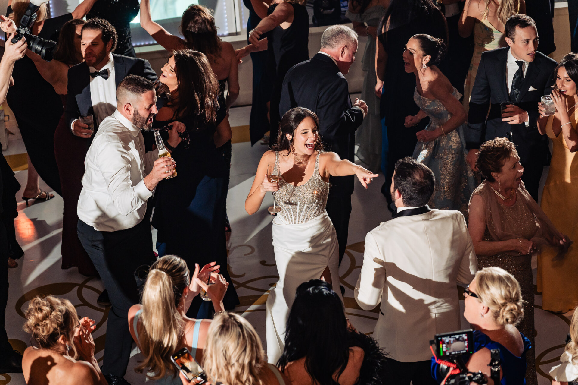 Bride dances with guests on the ballroom floor during the wedding reception, holding a drink and laughing as friends surround her.