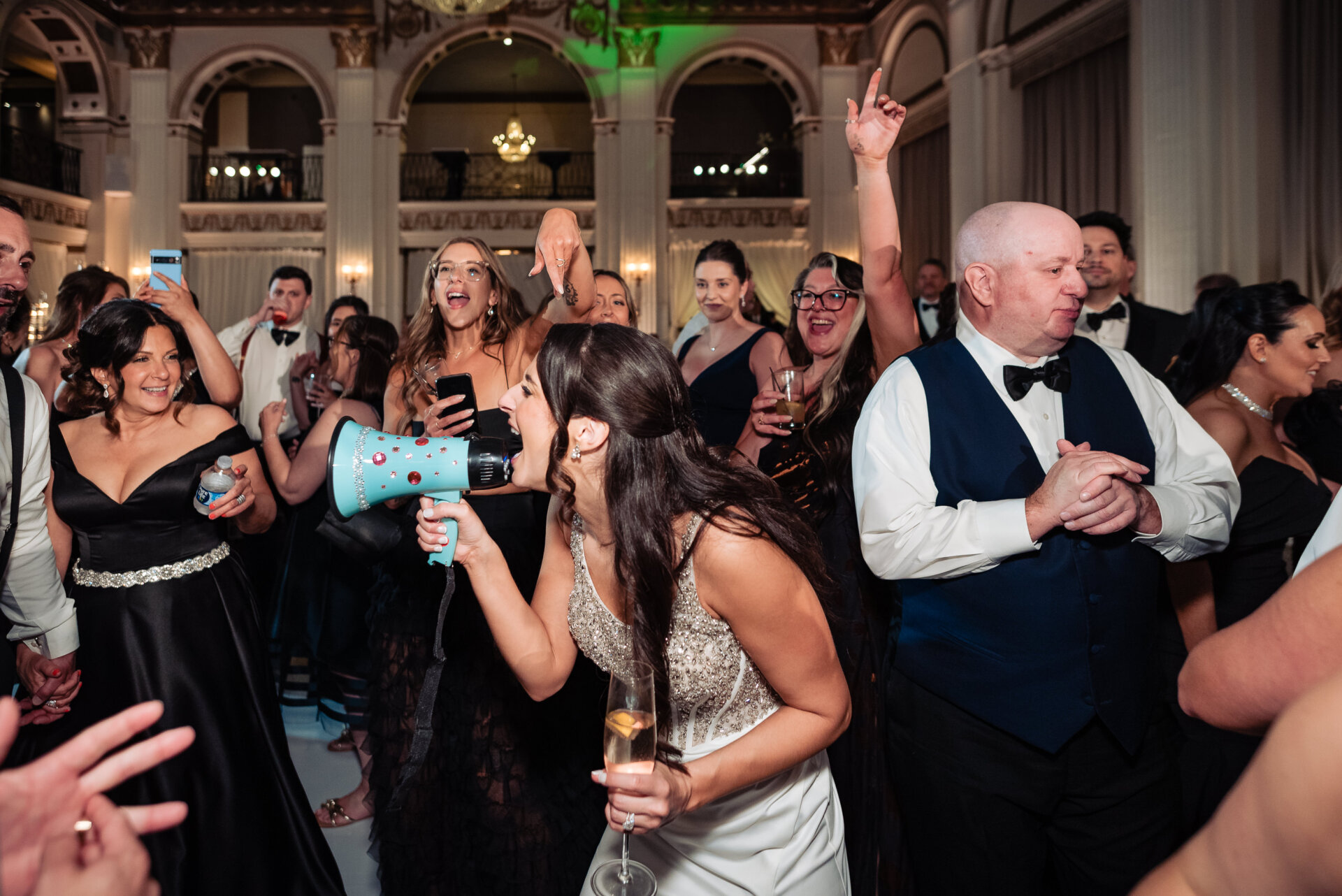 Bride leads the crowd in celebration, holding a megaphone and champagne glass while guests cheer on the dance floor at The Ben wedding reception.