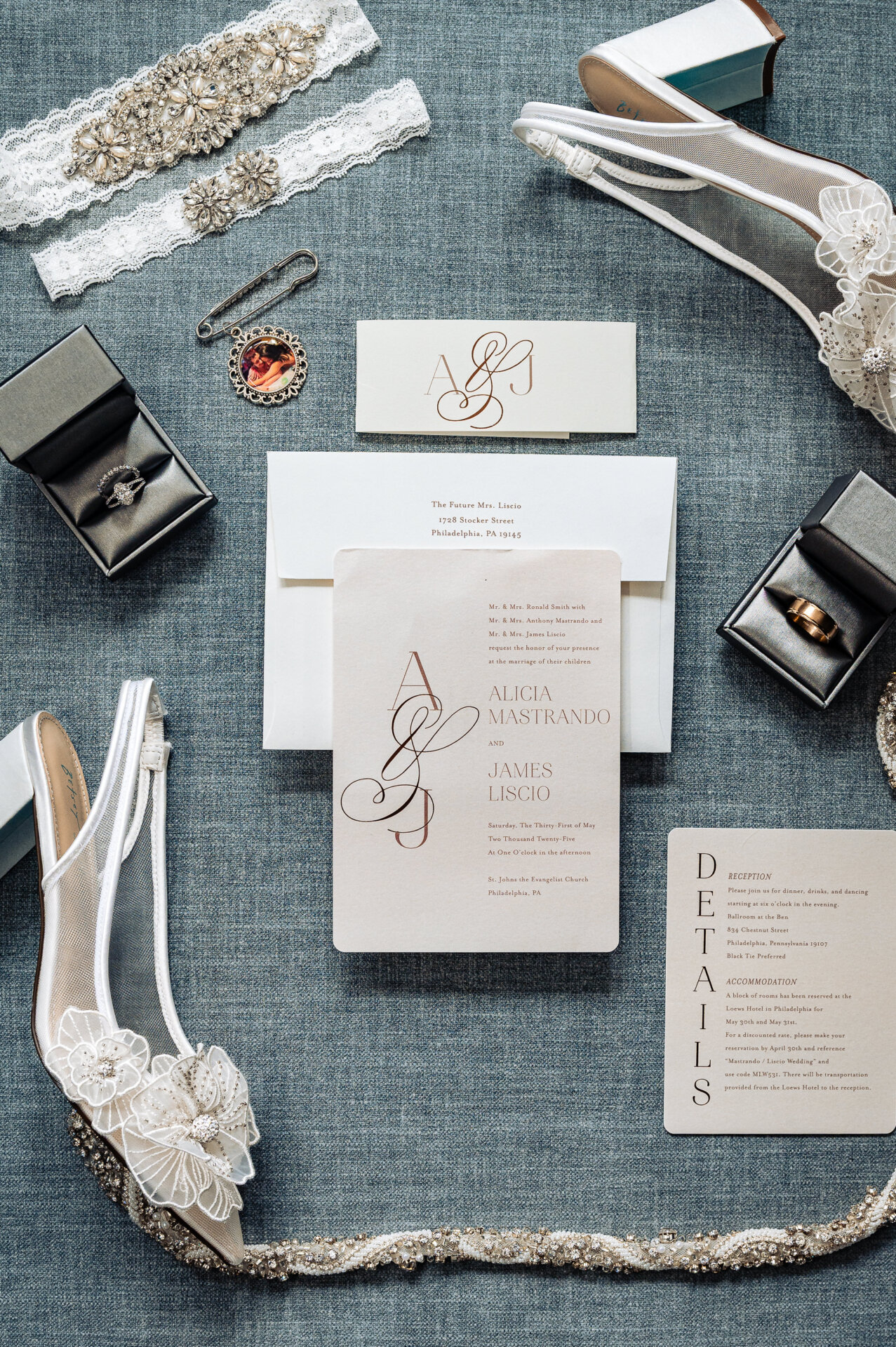 Wedding invitation suite with beige card, white shoes, rings, and decorative accessories laid out on gray fabric