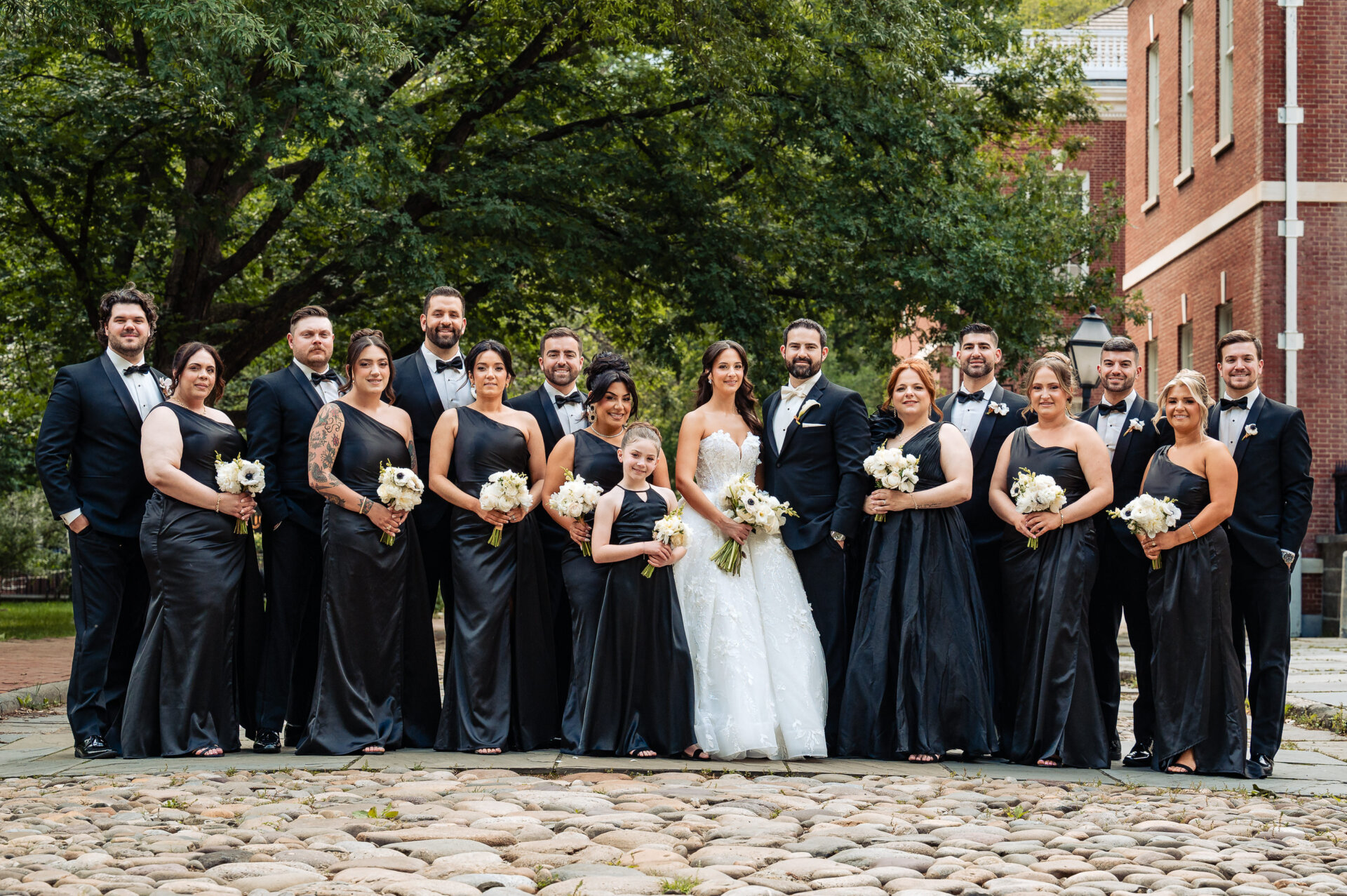 Bride and groom pose with their full wedding party outdoors, bridesmaids in black dresses and groomsmen in black tuxedos.