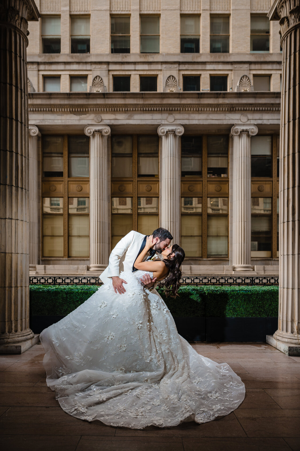 Groom kisses his wife as she leans slightly back, framed by three large marble columns before the wedding reception.