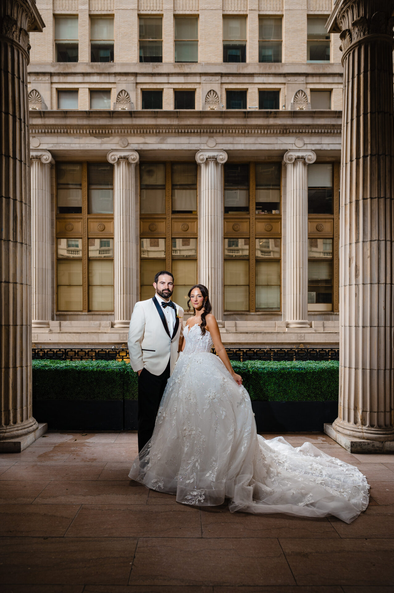 Bride and groom stand together between tall stone columns for a formal wedding portrait outside The Ben.