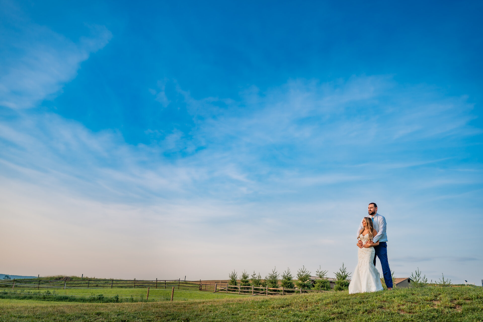 Couple standing together on a grassy hill with open countryside and blue sky during golden hour wedding portraits