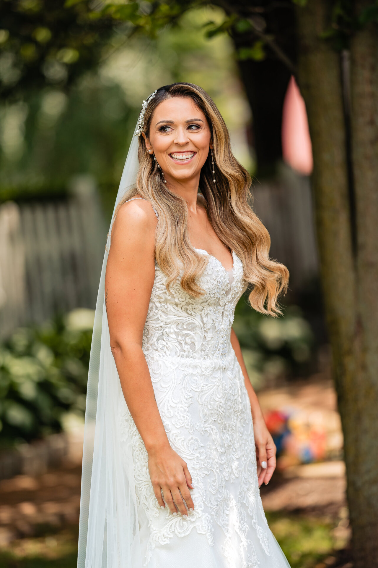 Bride portrait outdoors, smiling in her lace wedding dress with veil and natural greenery behind her