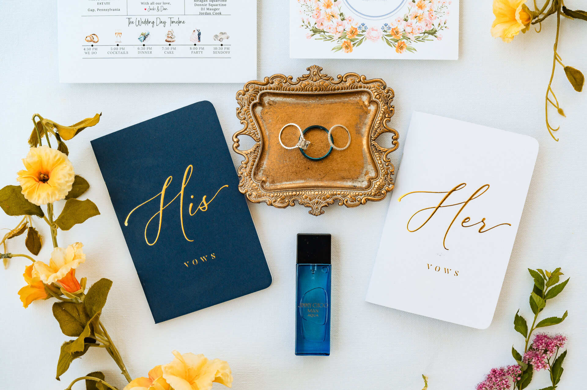 Flat lay of wedding details featuring rings, his and her vow books, and floral accents.