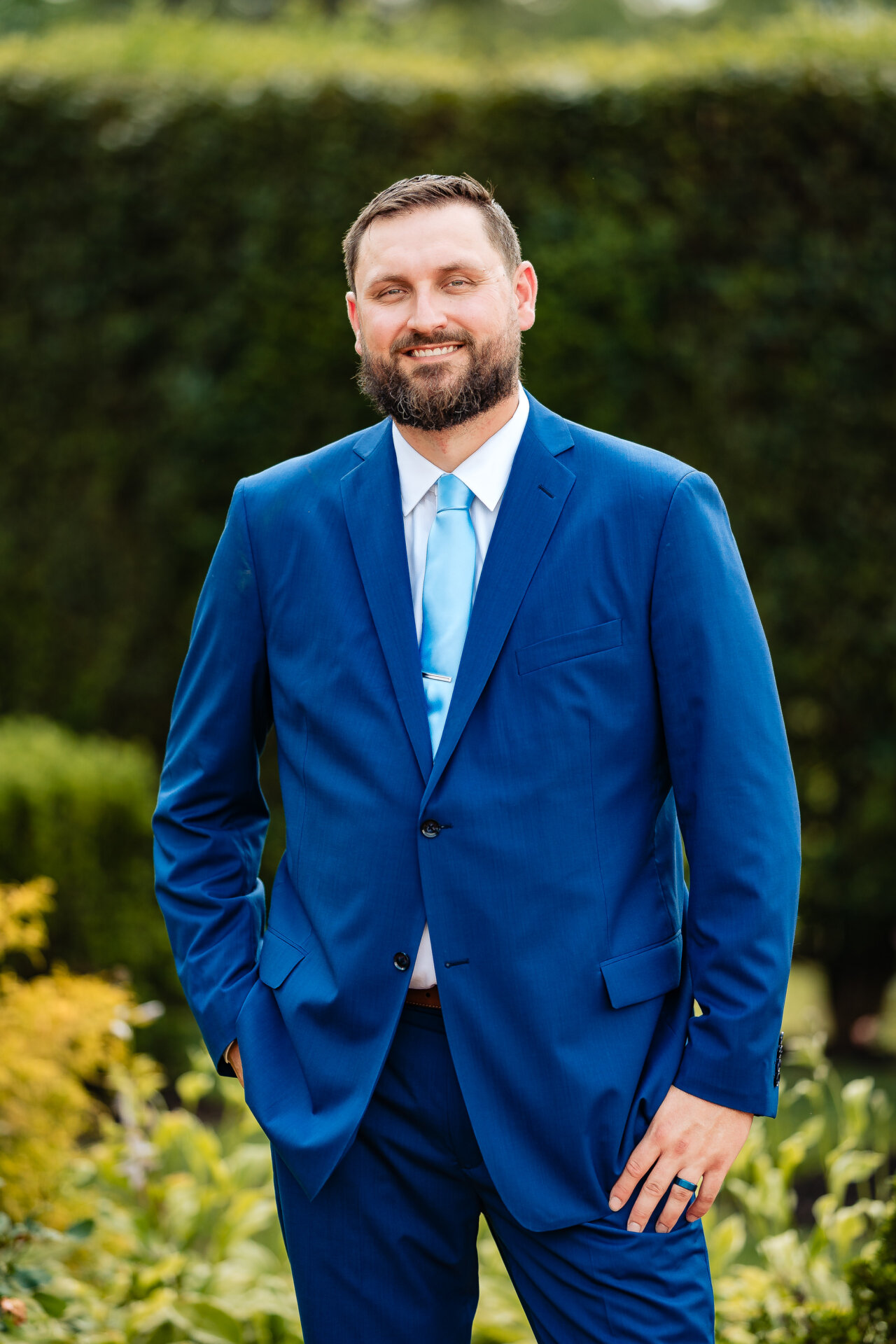 Groom portrait in a blue suit standing outdoors with greenery in the background