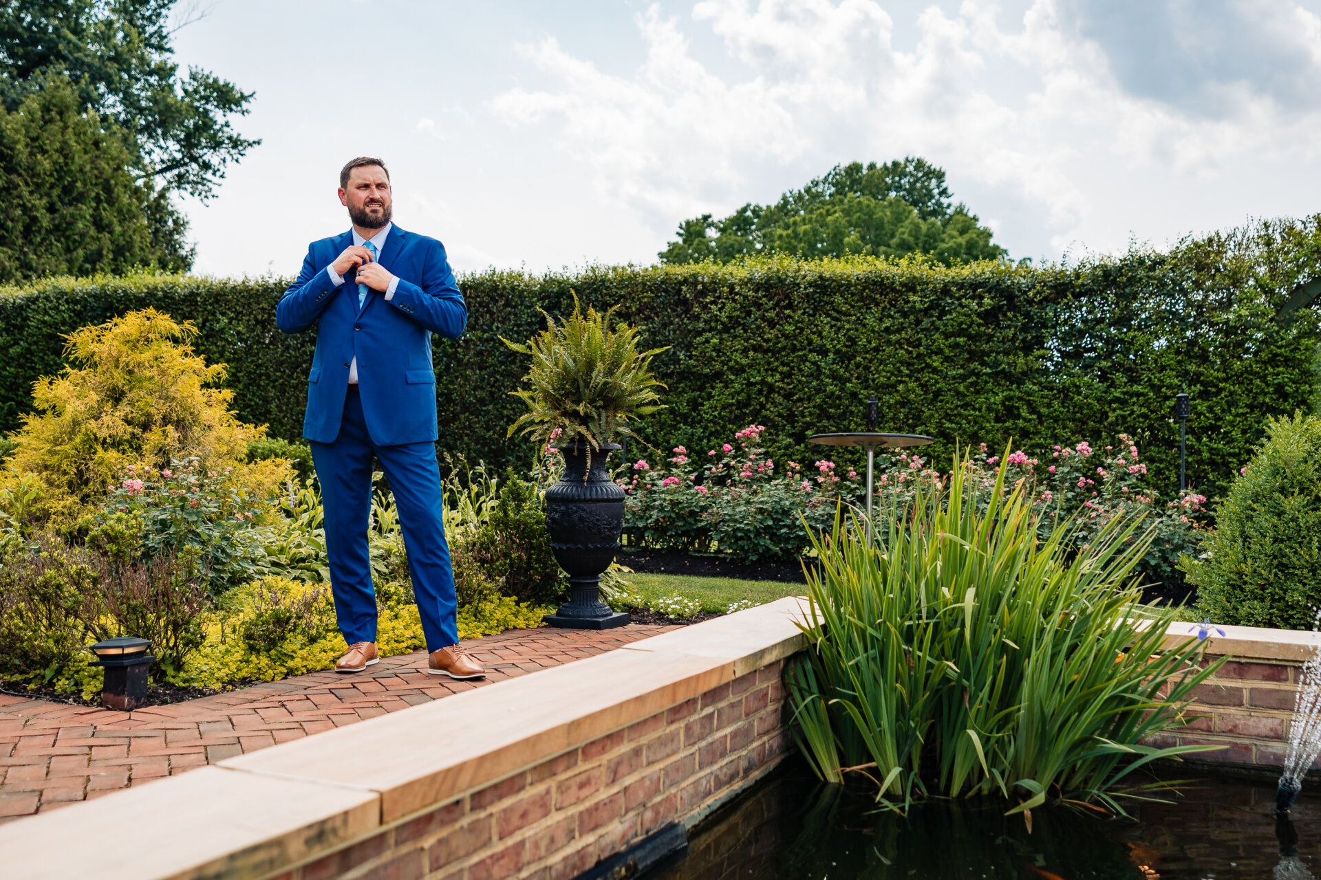 Groom adjusting his suit tie while standing beside a garden pond before the ceremony