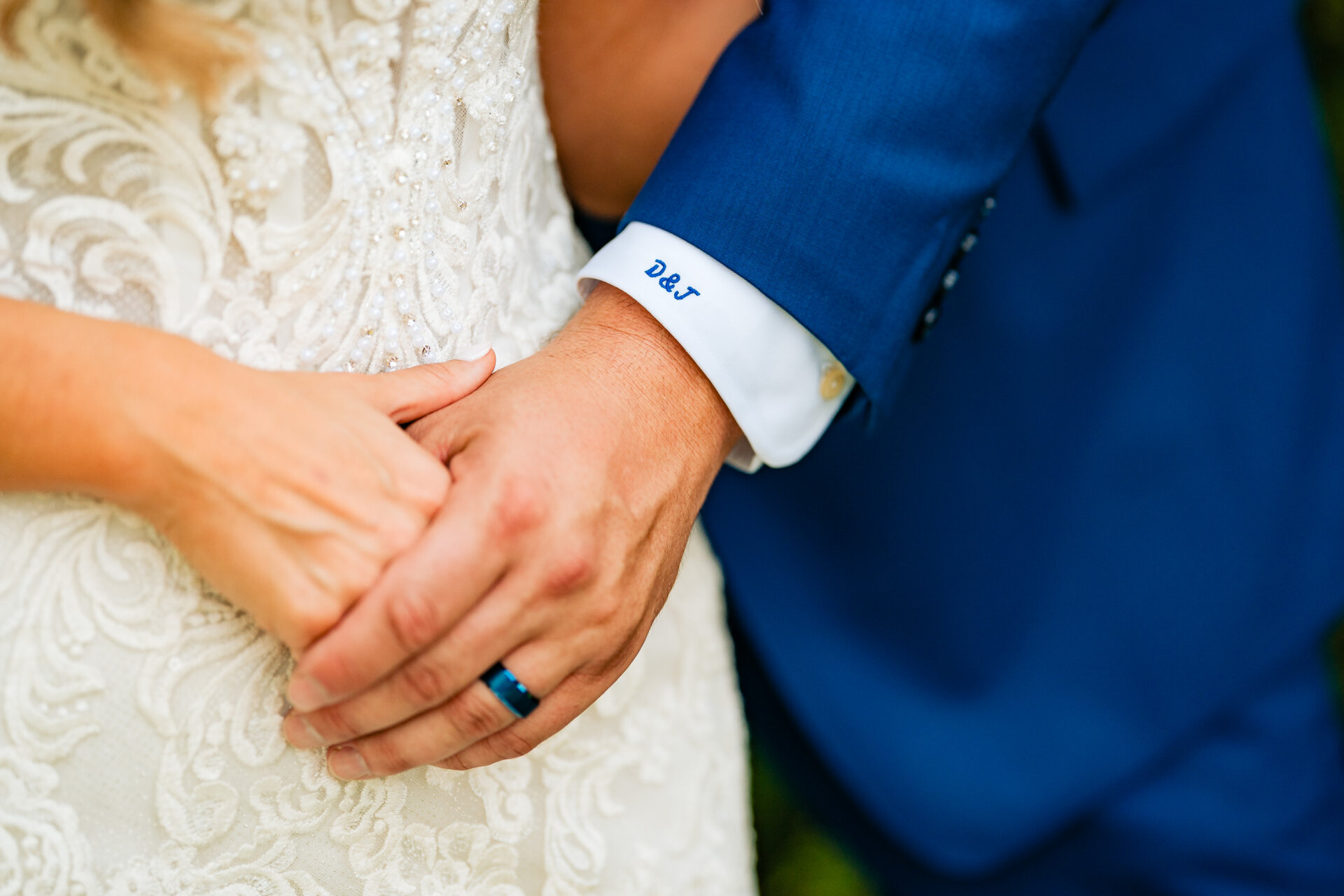 Close-up of bride and groom holding hands, showing embroidered initials on the groom’s cuff and wedding rings