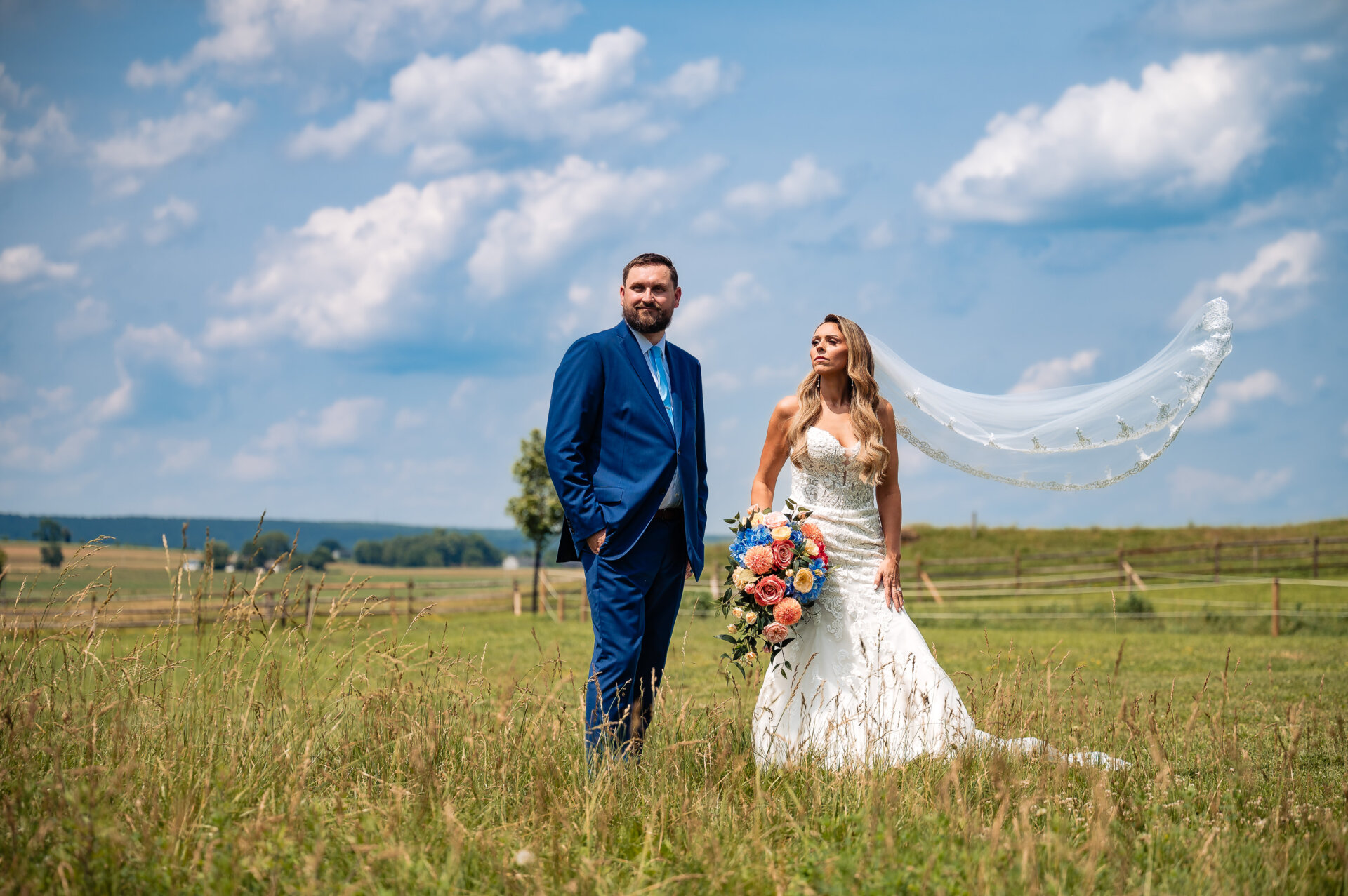Bride and groom stand together in an open field as the bride’s veil flows in the wind.