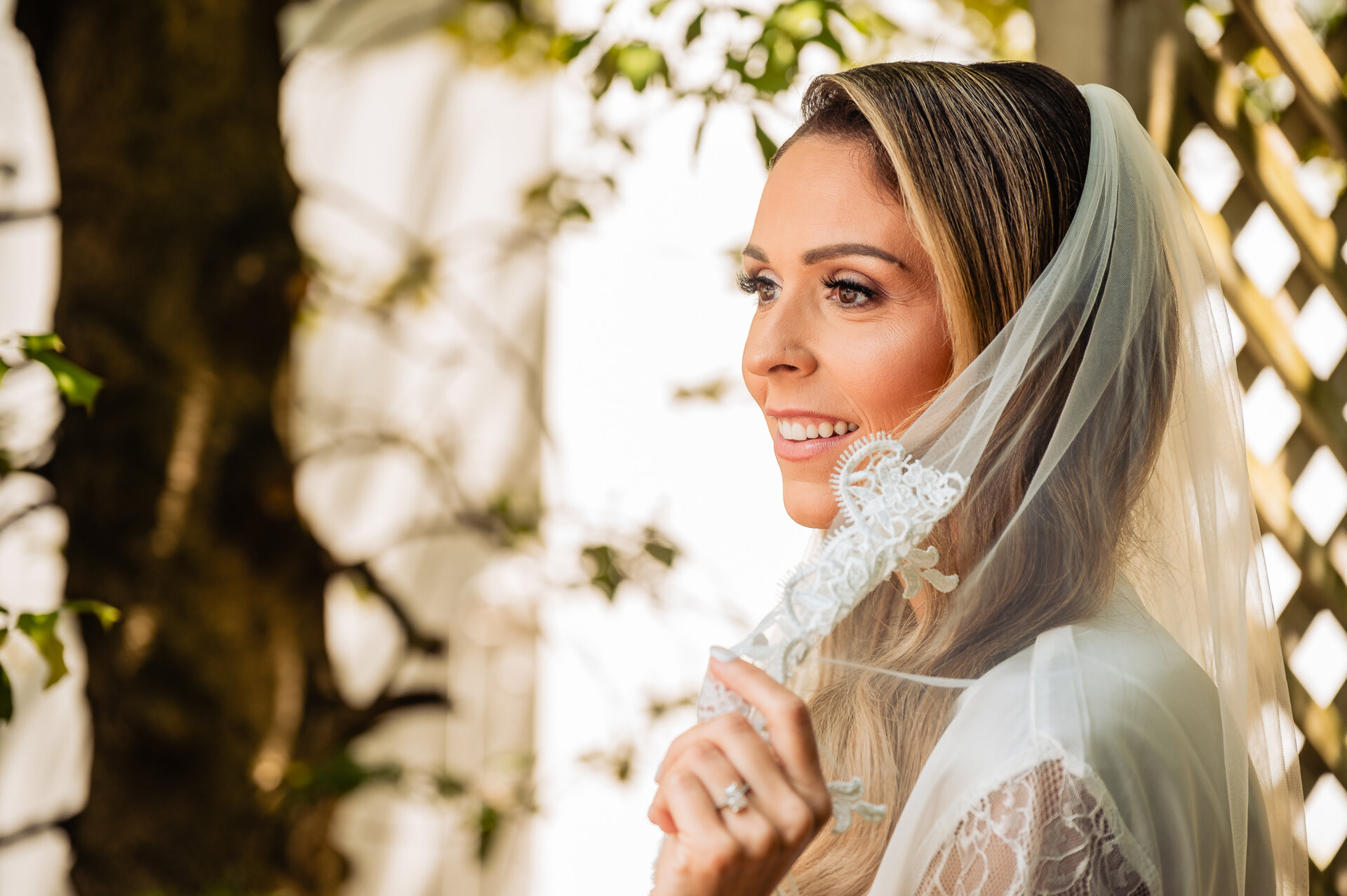 Bride in her wedding veil smiles and looks off-camera while holding the lace edge of her veil during a portrait session outdoors.