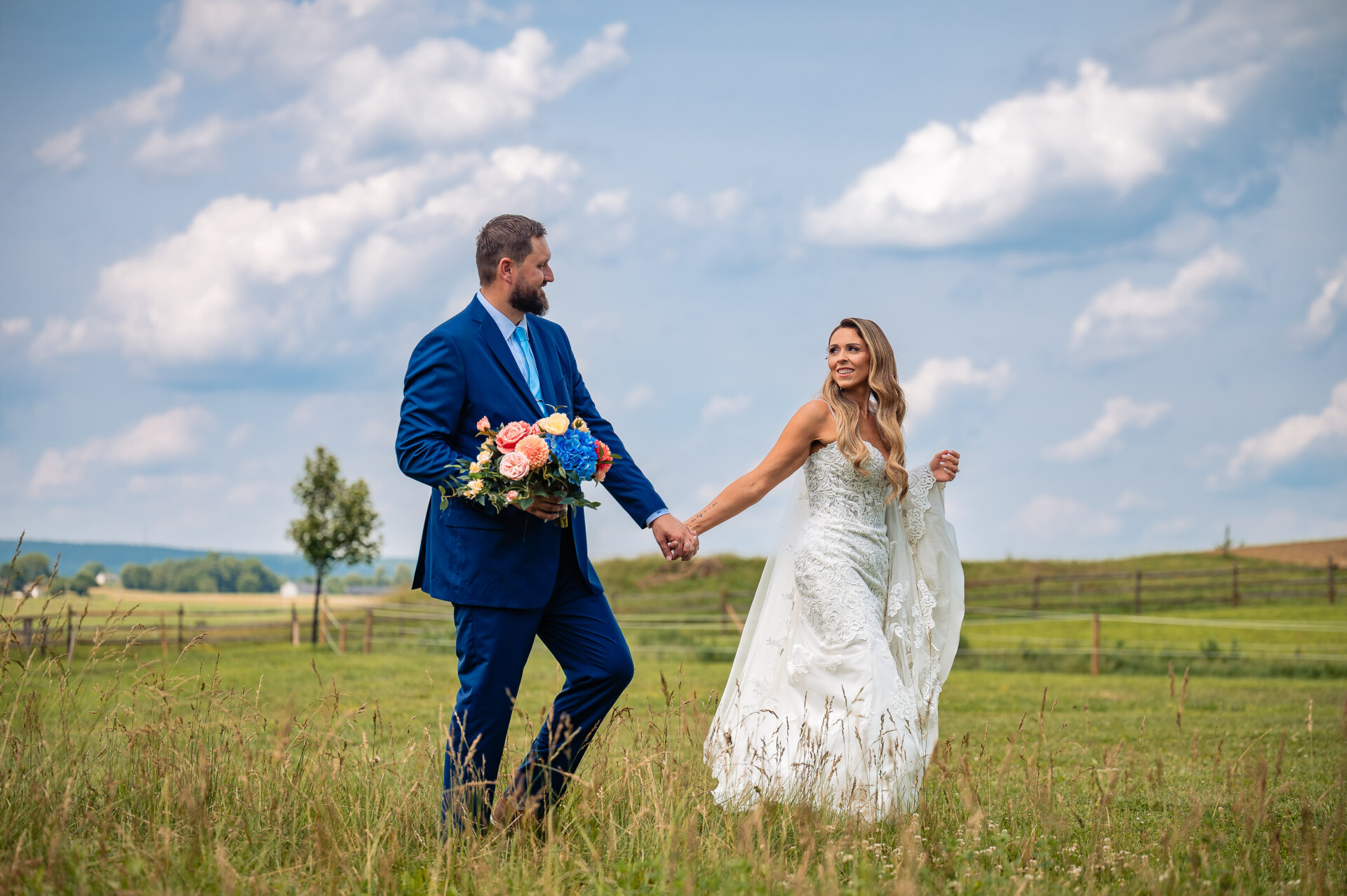 Bride and groom walking hand in hand through a grassy field under a bright blue sky.