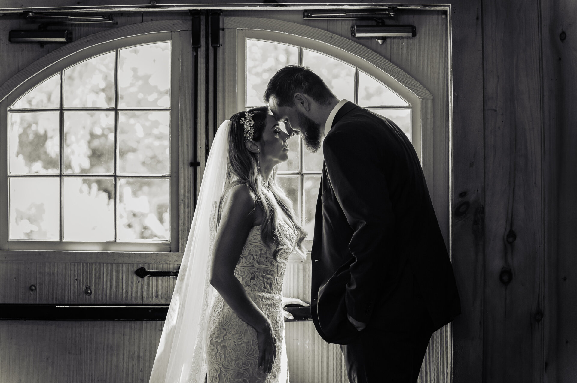 Black and white portrait of bride and groom touching foreheads by a window at the white chimney estate