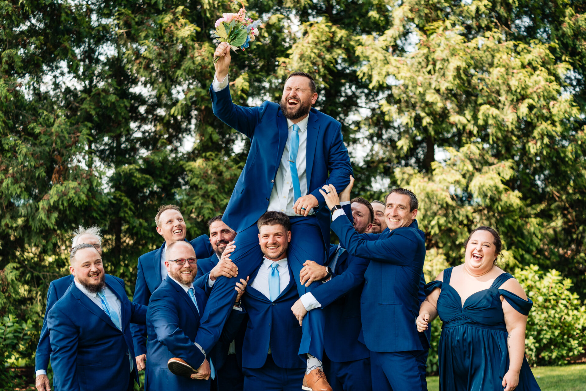 Groom lifted by his wedding party outdoors while holding a bouquet and laughing