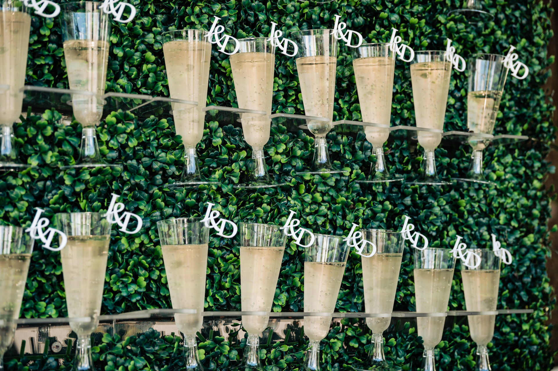 Champagne wall display with personalised “J & D” drink tags set against a lush greenery backdrop at the wedding reception