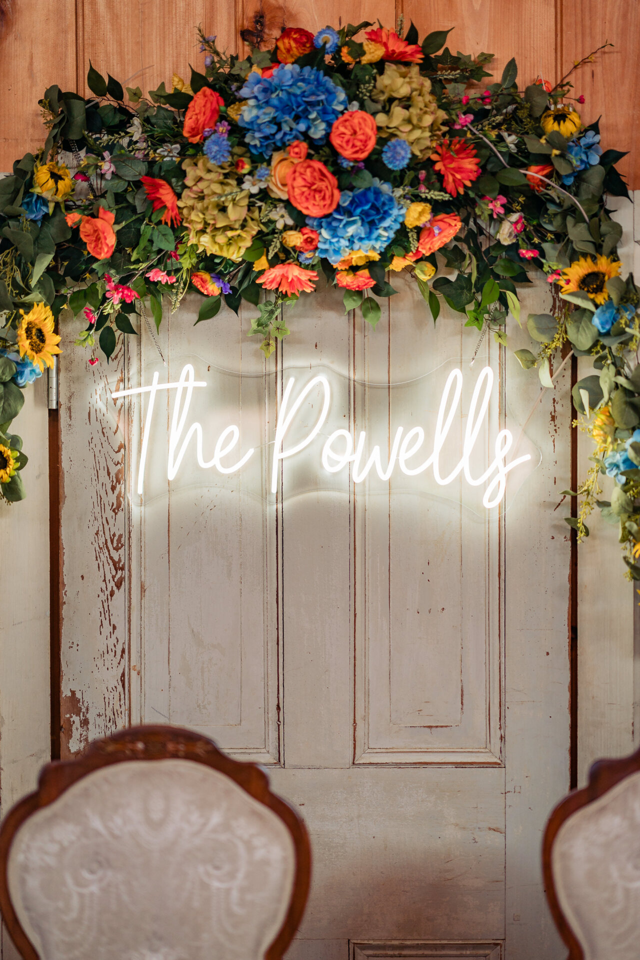 Wedding reception floral backdrop with a neon sign reading “The Powells”