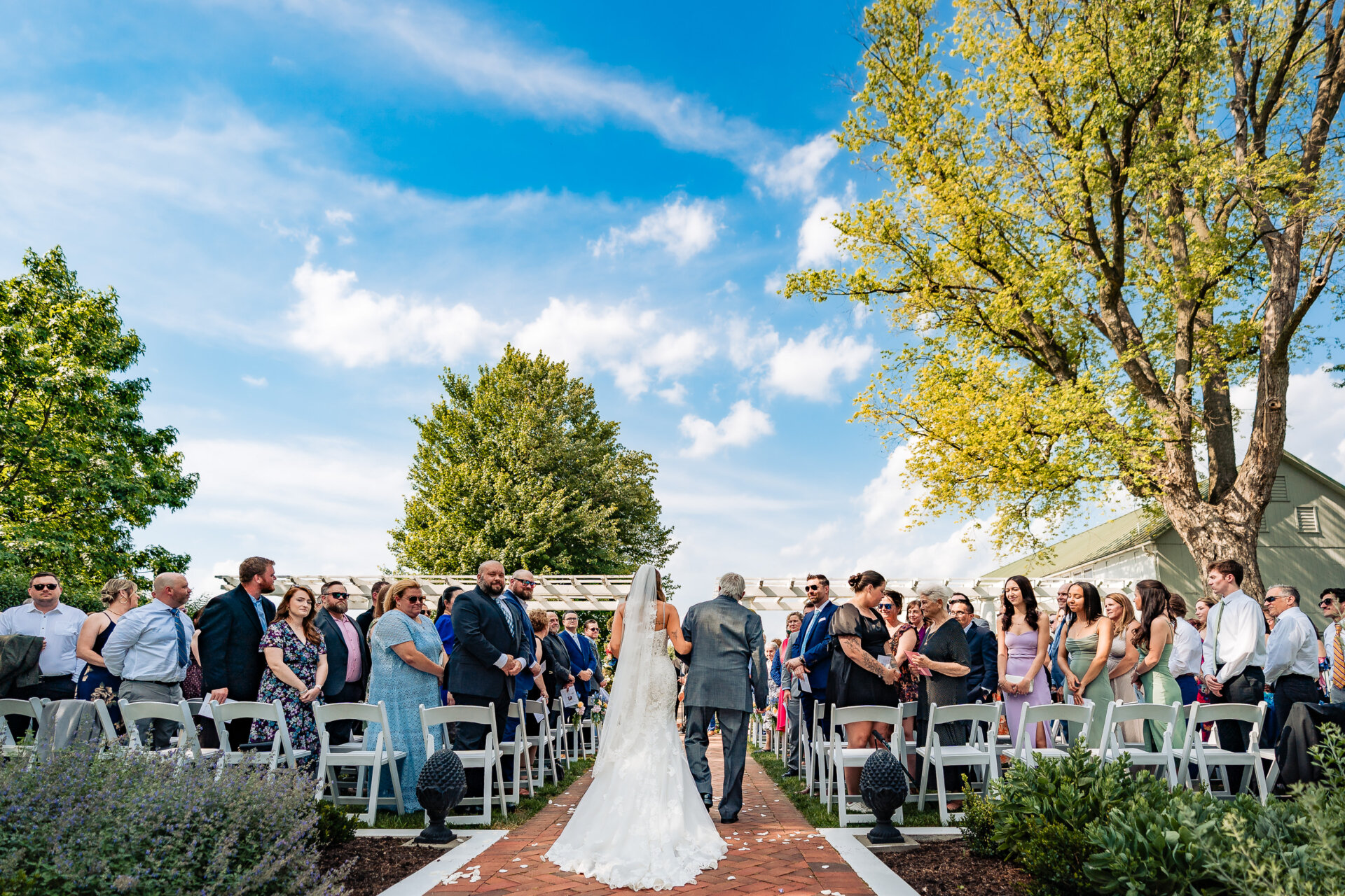 Bride walking down the aisle with her father during an outdoor wedding ceremony, guests standing on both sides