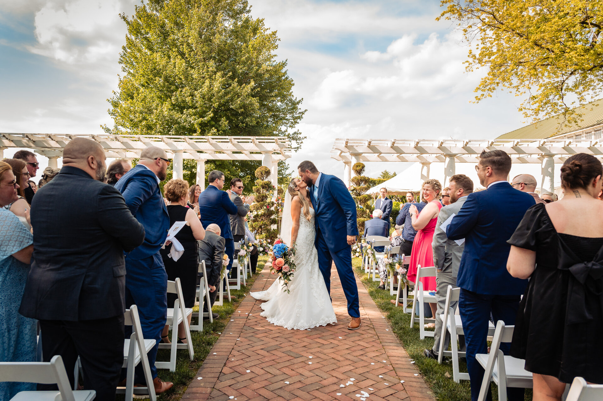 Bride and groom kiss at the altar under a pergola as guests applaud during the ceremony.
