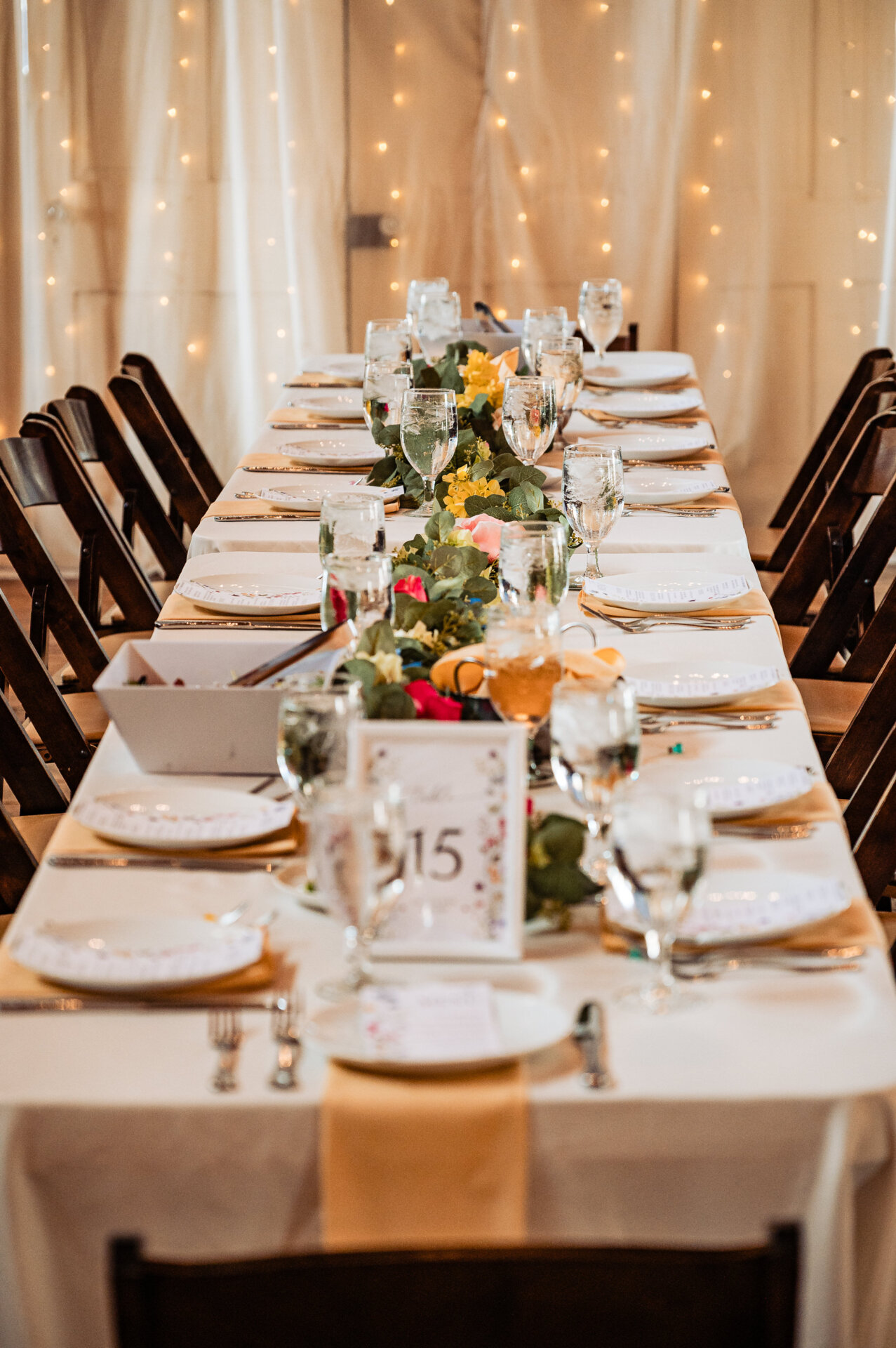 Long reception table styled with floral greenery, place settings, and soft string lighting.