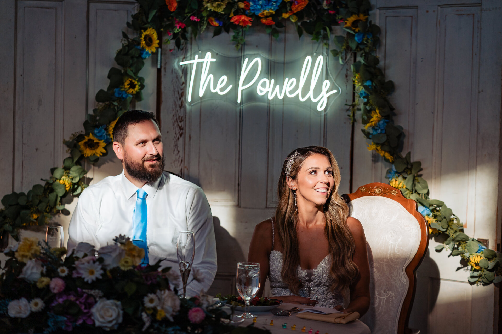 Bride and groom seated at their sweetheart table beneath a neon “The Powells” sign during the wedding reception