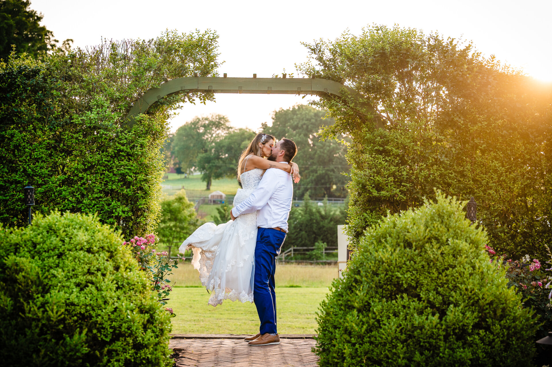 Bride and groom sharing a kiss beneath a garden arch framed by greenery at sunset