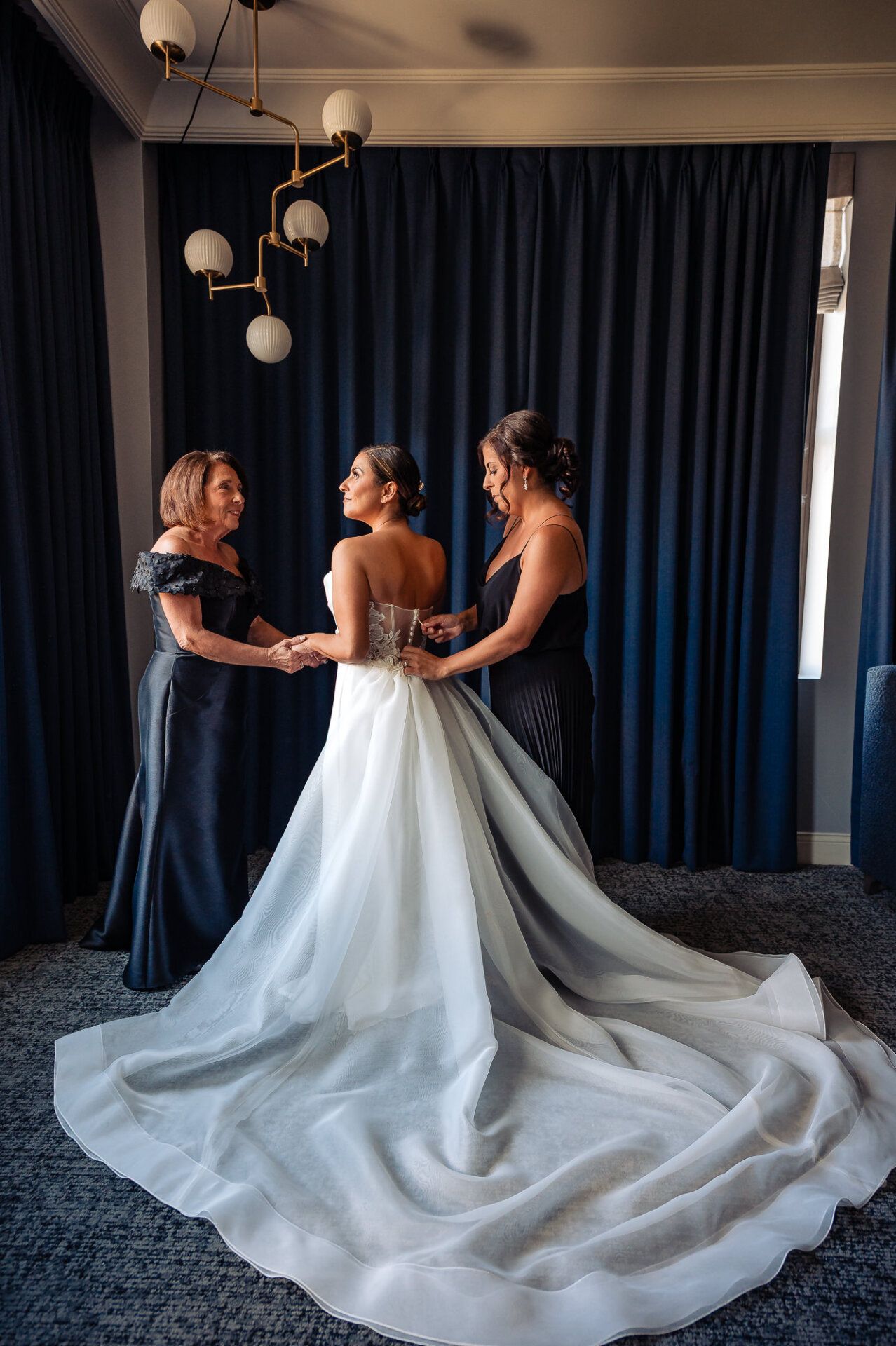 Bride in strapless ballgown with dramatic flowing train being helped by mother and bridesmaid in navy dresses under modern globe chandelier