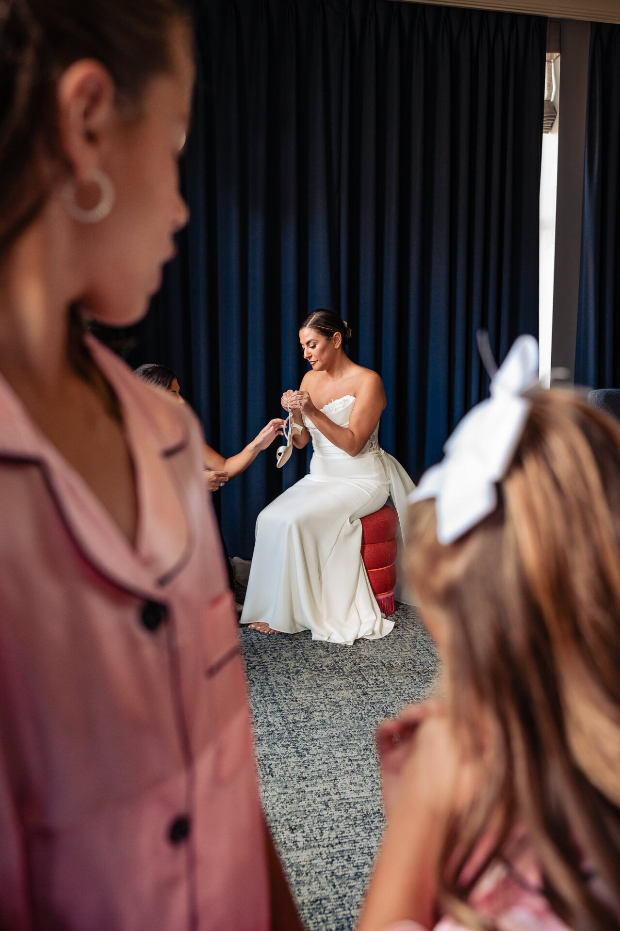 Bride in white strapless gown getting ready with bridesmaids in pink attire against navy curtain backdrop