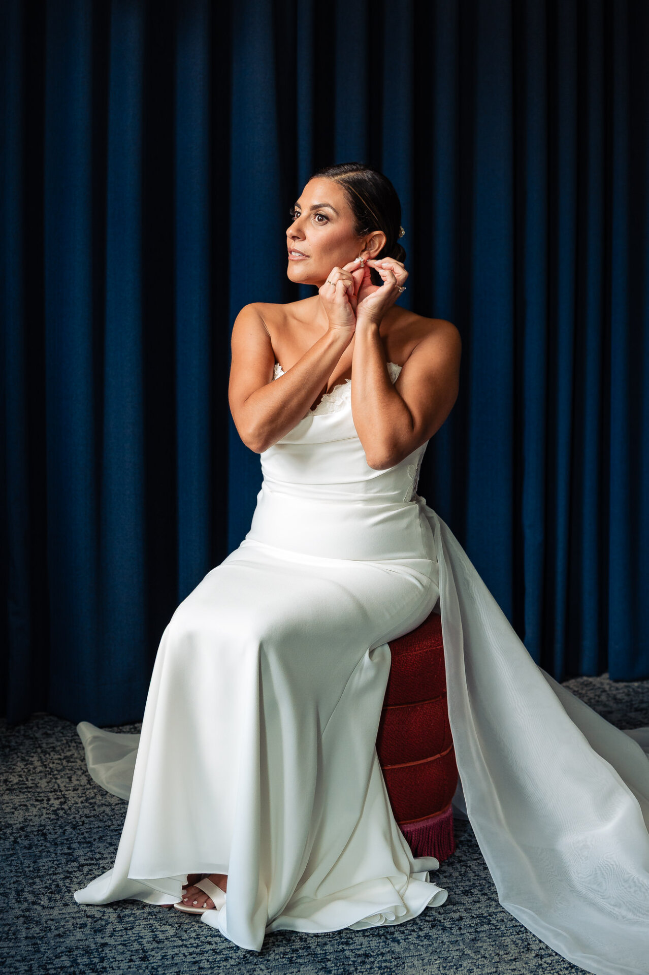 Bride adjusting pearl earring while seated on red ottoman in white strapless gown against navy curtain