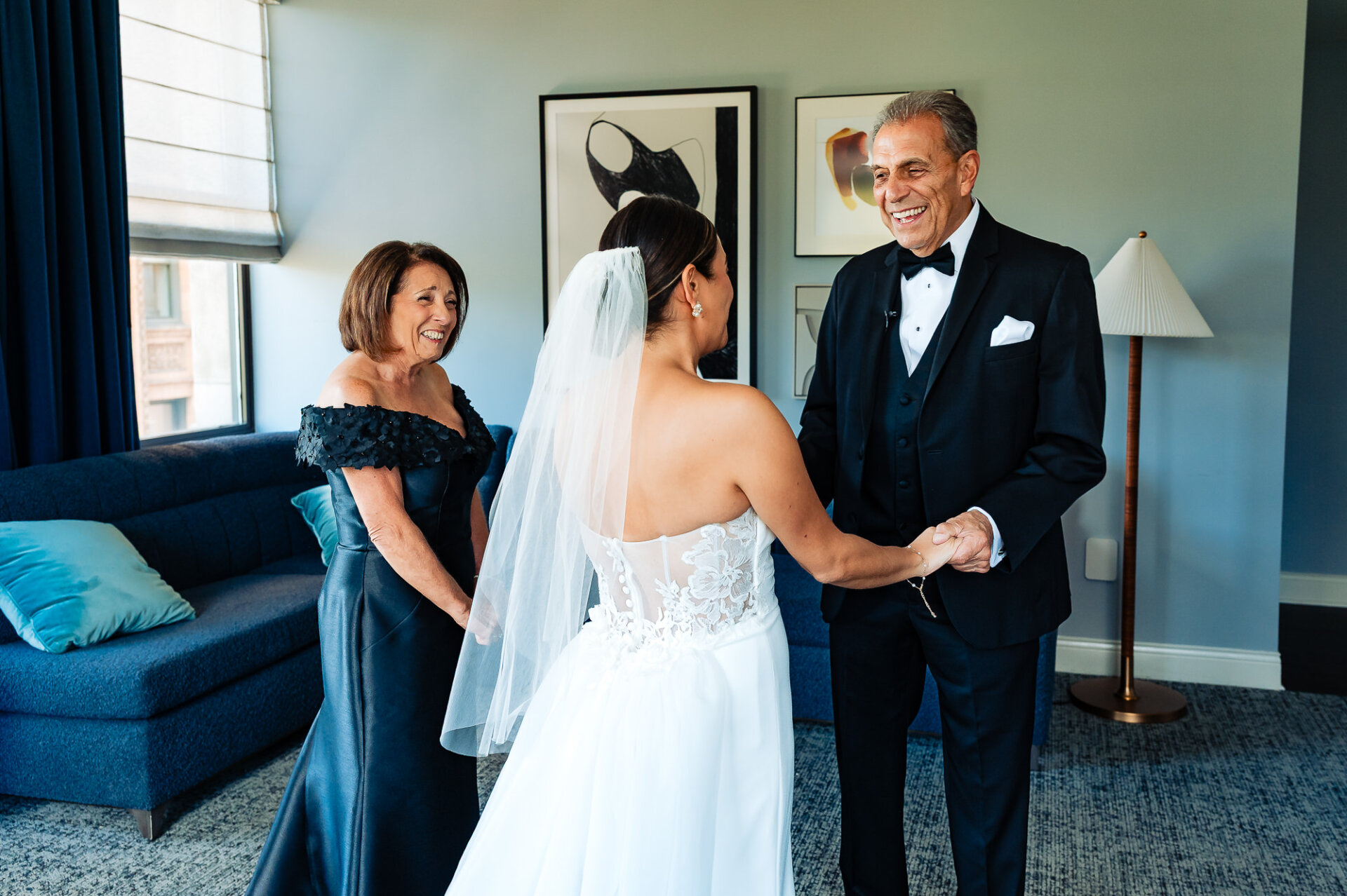 Bride in veil sharing emotional first look moment with parents in formal attire in blue hotel suite