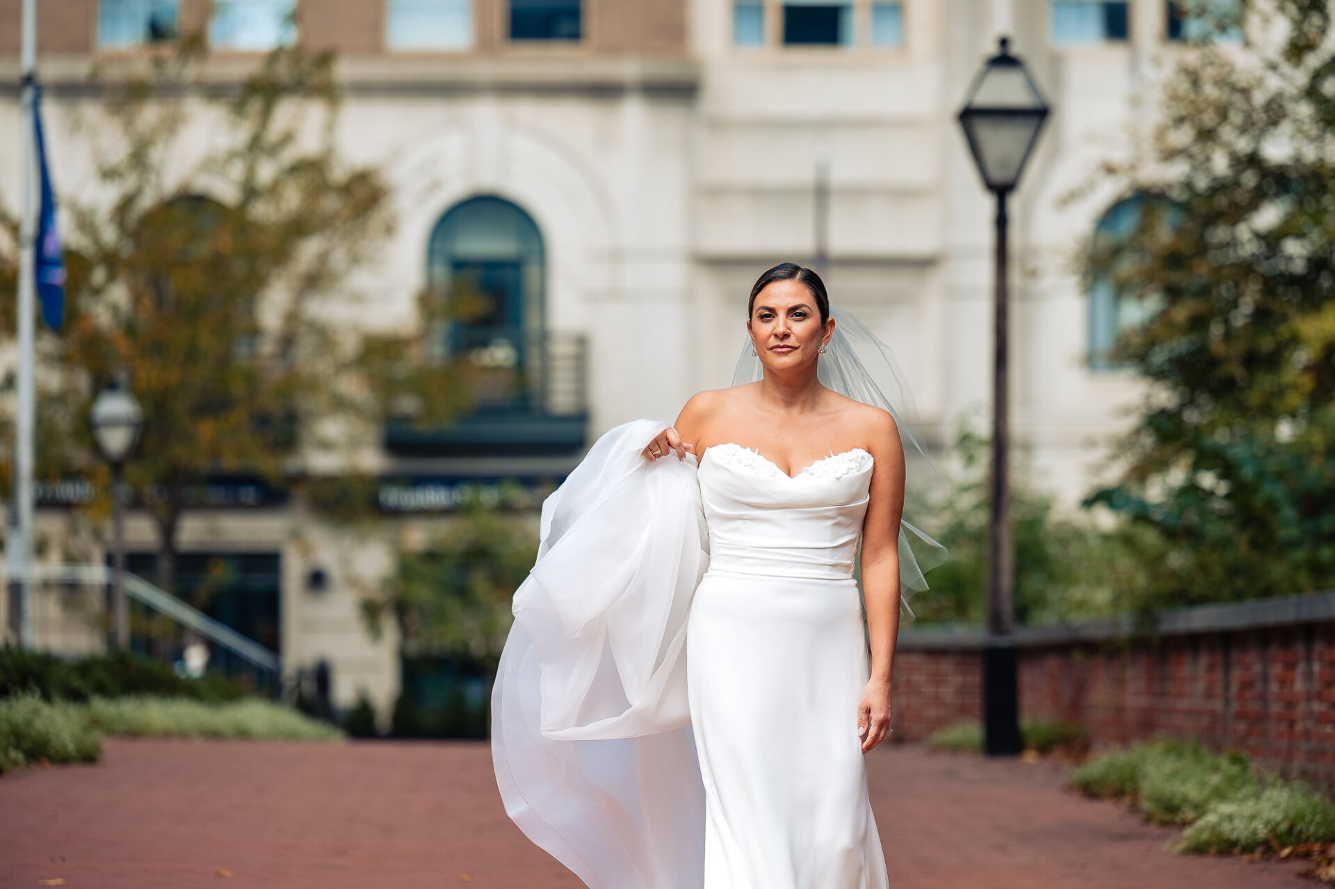 Bride in strapless white gown holding veil in breeze on brick walkway with historic building backdrop