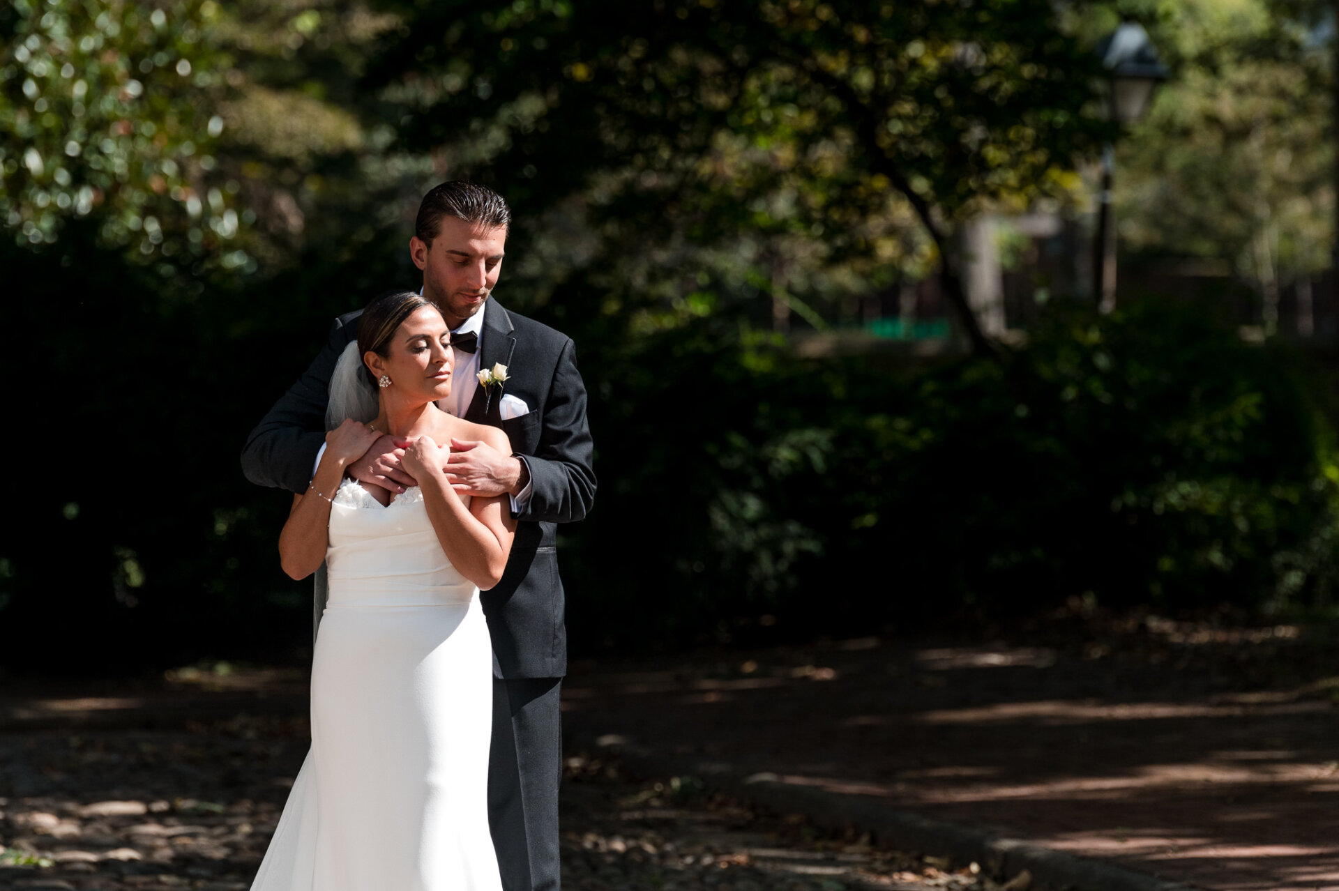 Groom embracing bride from behind in sunlit garden, both in formal wedding attire