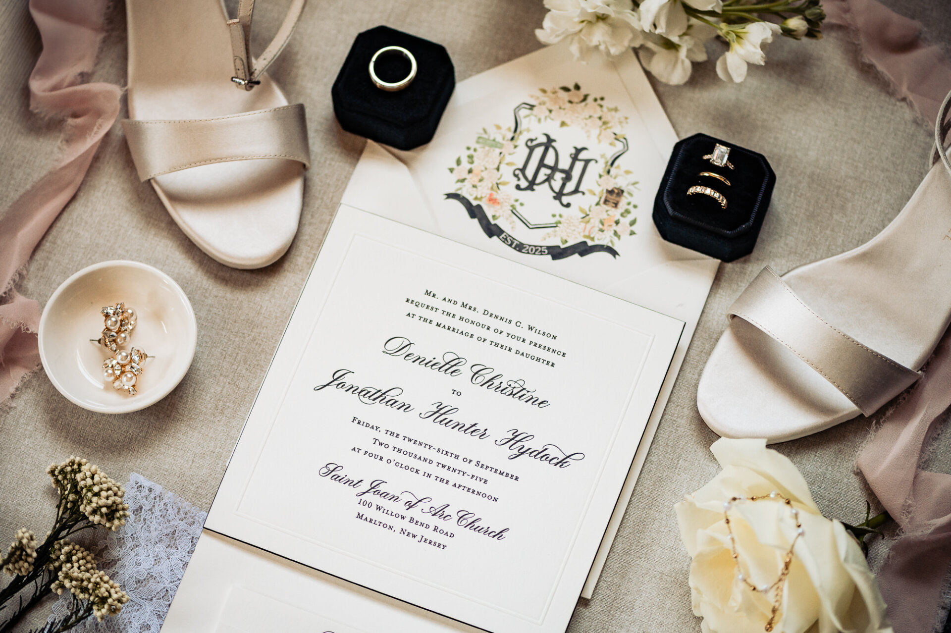 Wedding flat lay with invitation, engagement and wedding rings in black velvet boxes, pearl earrings, champagne heels, and white flowers