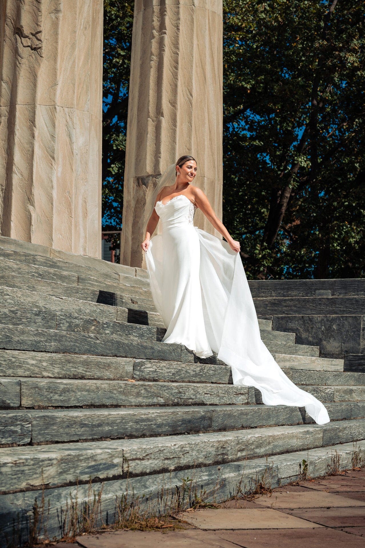 Bride descending stone steps in flowing white gown with dramatic train between tall marble columns