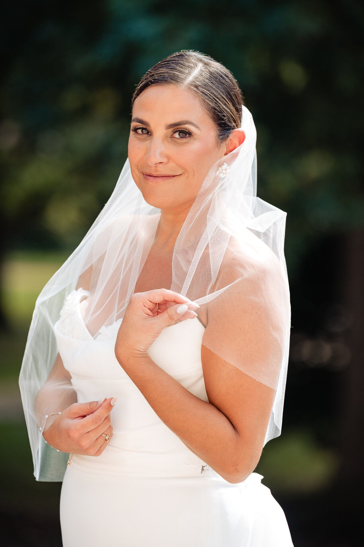 Bride smiling at camera wearing white strapless gown and flowing veil with natural outdoor background
