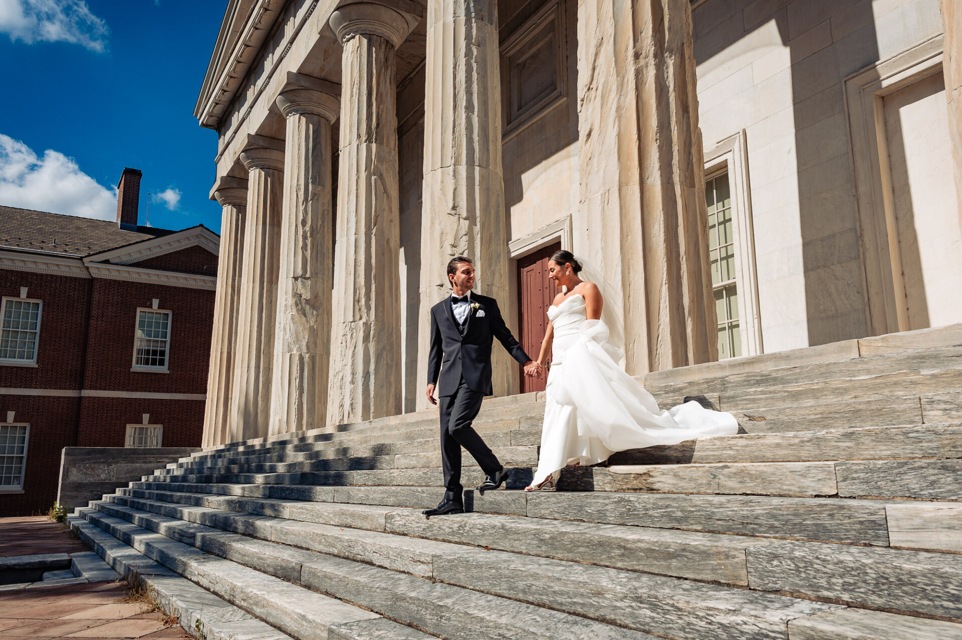 Newlywed couple holding hands on steps of marble building, bride in white gown and groom in black tuxedo