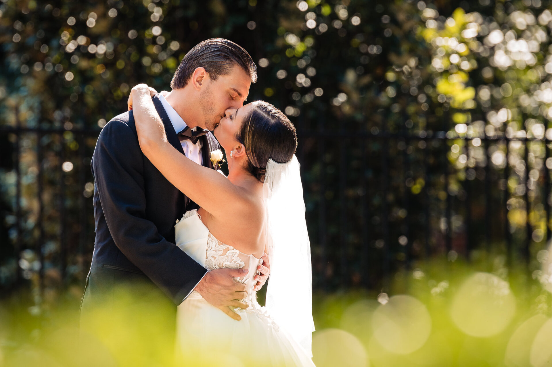 Bride and groom kissing in garden with sunlit bokeh background and iron fence