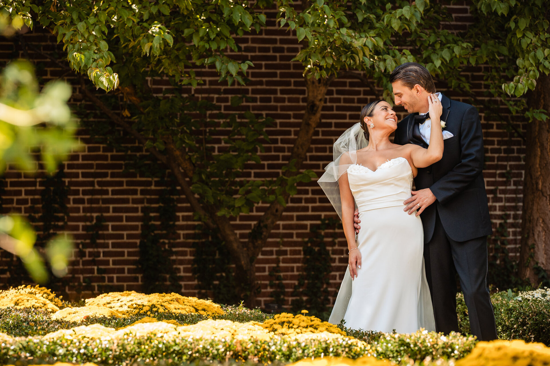 Newlyweds embracing in garden with vibrant yellow mums and brick wall background