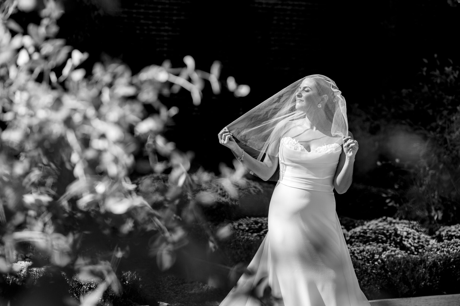 Black and white photo of bride holding veil in wind wearing strapless gown in garden setting