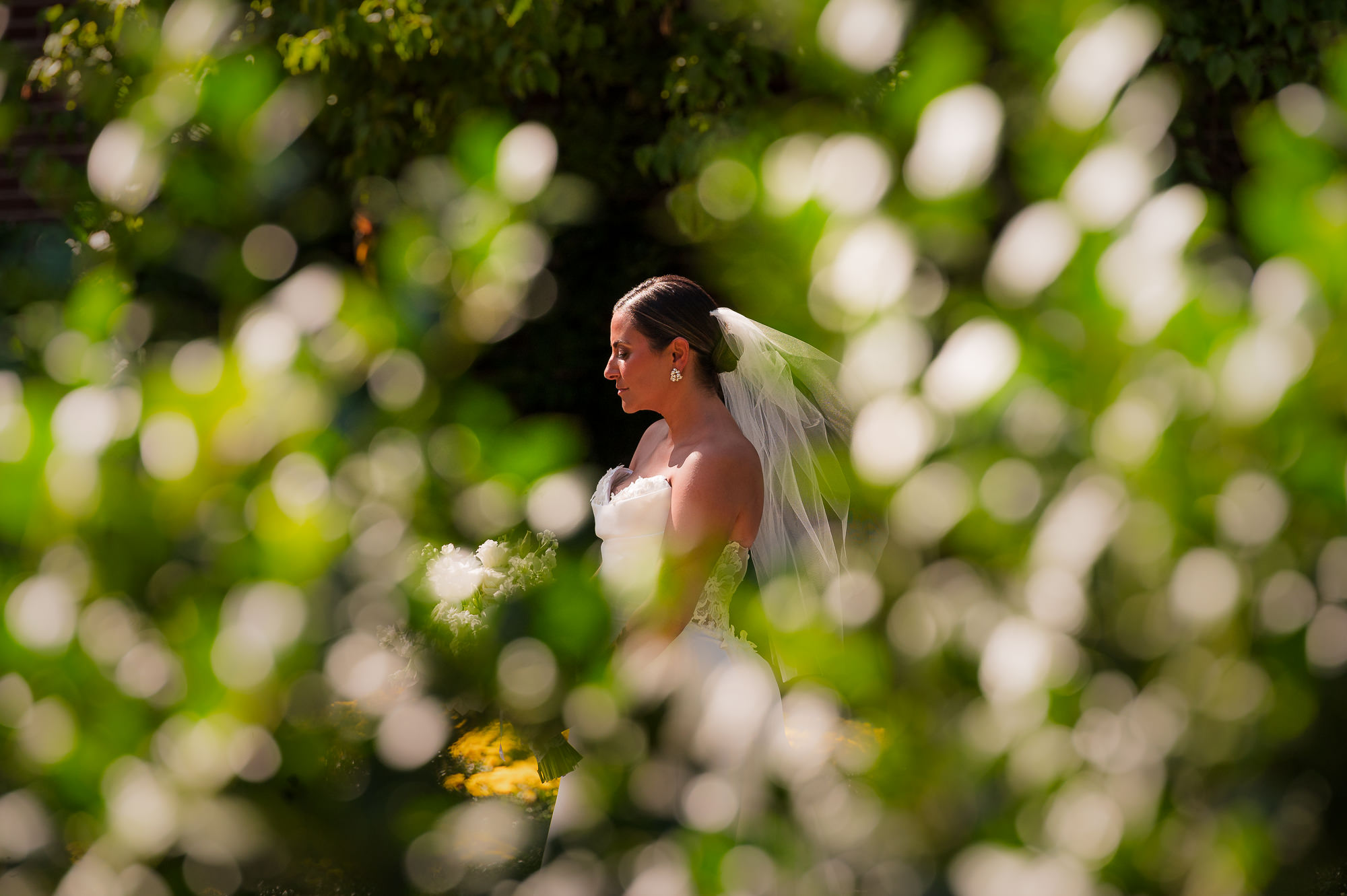 Bride in profile holding white bouquet with veil, framed by natural foliage bokeh in foreground