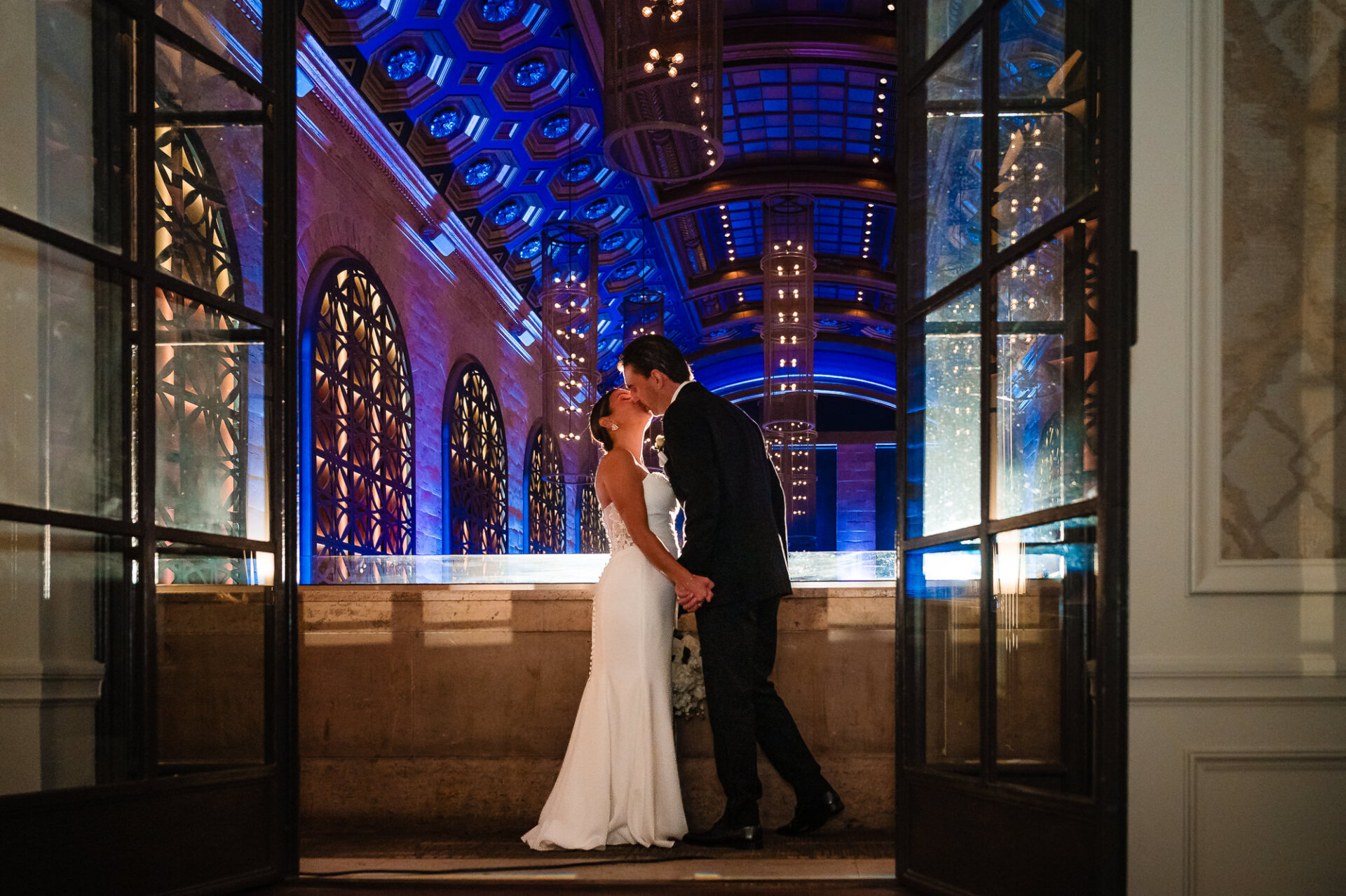 Bride and groom kissing in doorway framed by ornate venue with blue illuminated coffered ceiling