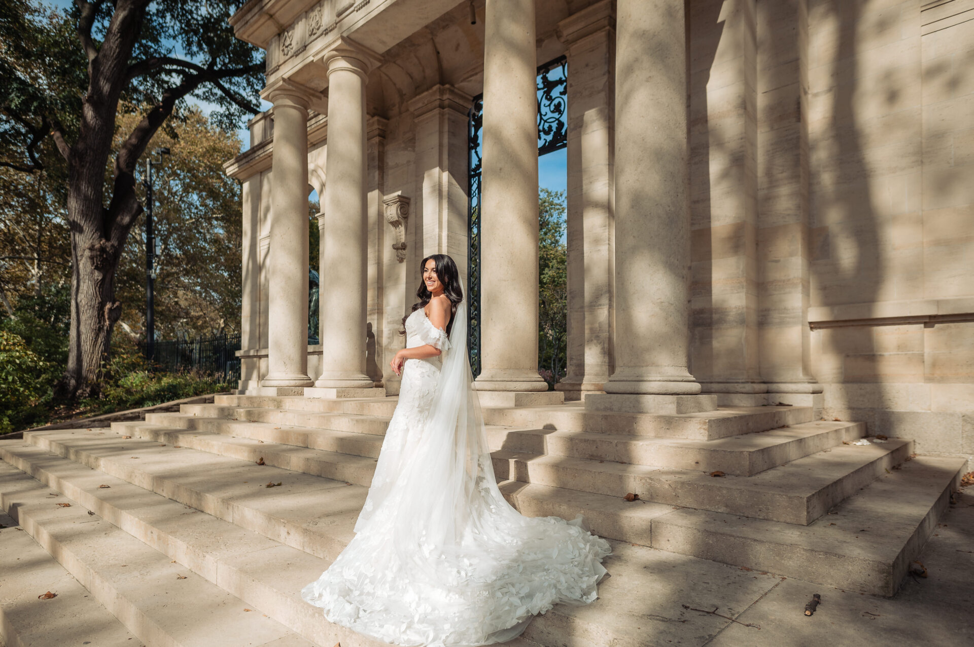 Bride in off-shoulder lace gown with cathedral train standing on marble steps among tall neoclassical columns in dappled sunlight