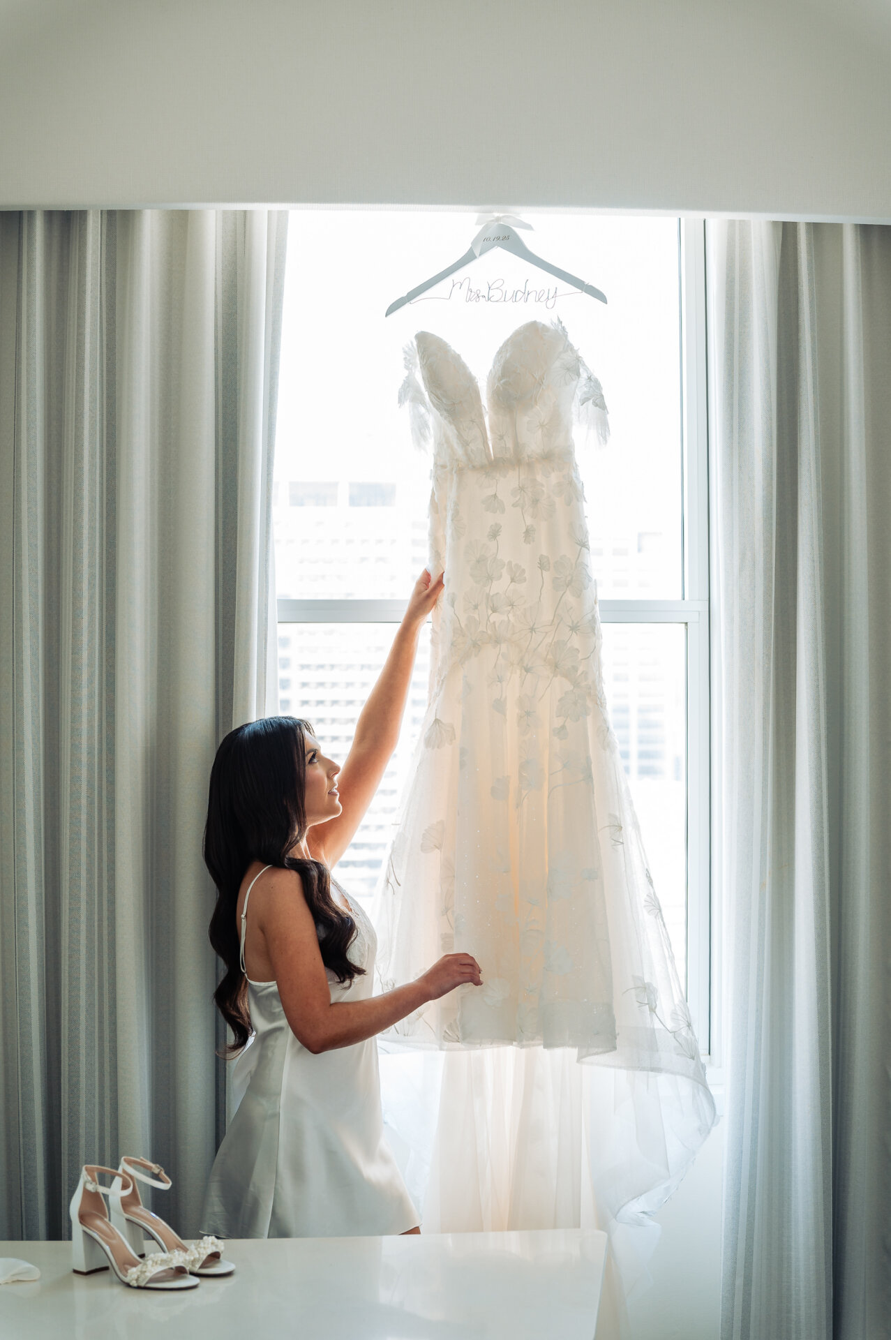 Bride in white slip dress admiring lace wedding gown with floral details hanging in bright hotel room window with pearl-embellished heels in foreground
