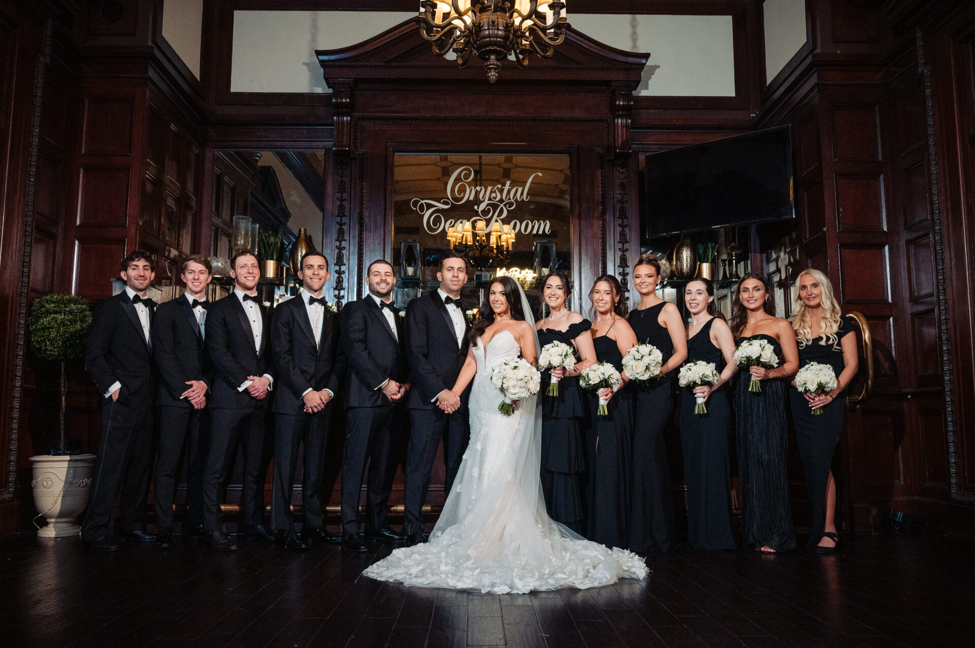 A full wedding party posing together inside the Crystal Tea Room, featuring dark wood paneling and a large mirror.