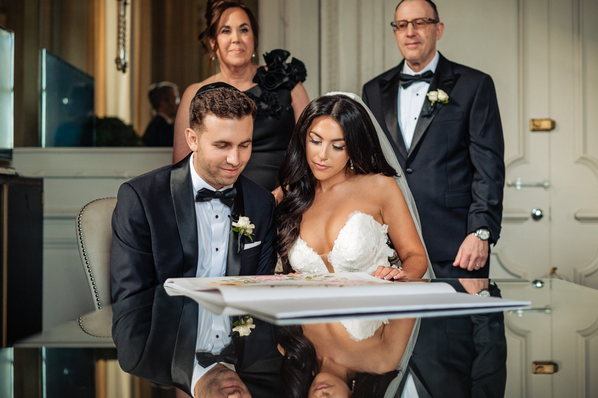 A rabbi or officiant points to a document while the bride and groom sit at a reflective table during a Ketubah signing.
