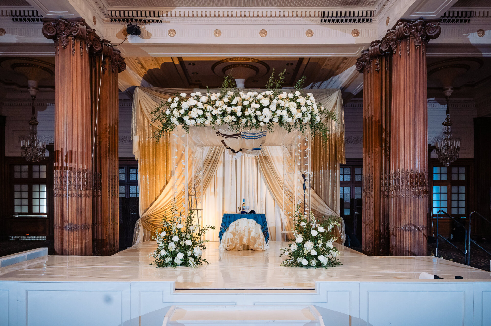 **Alt text:** Wedding ceremony chuppah adorned with white florals and draped fabric on a raised stage inside the Crystal Tea Room ballroom