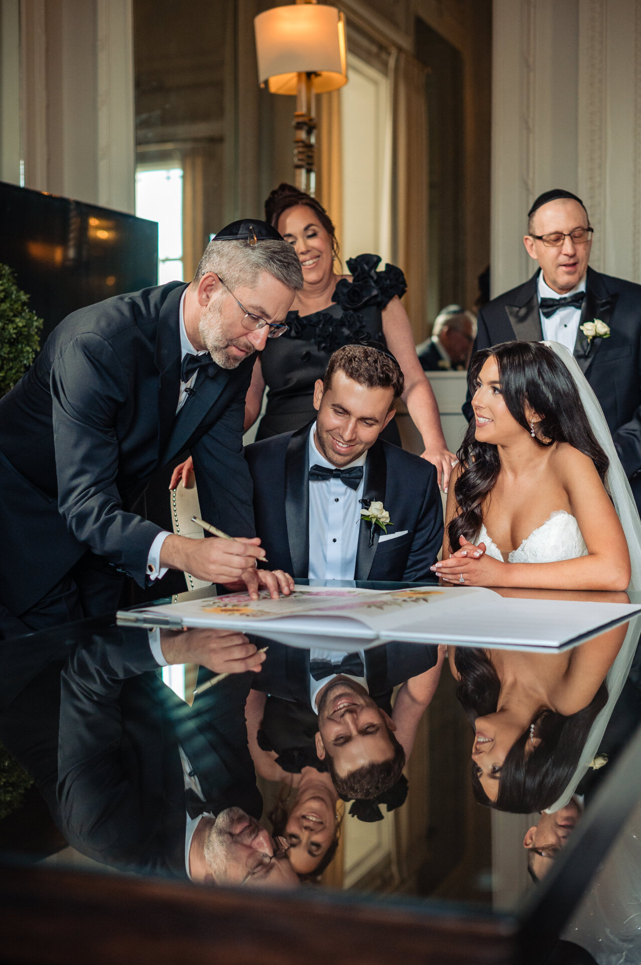 A rabbi or officiant points to a document while the bride and groom sit at a reflective table during a Ketubah signing.
